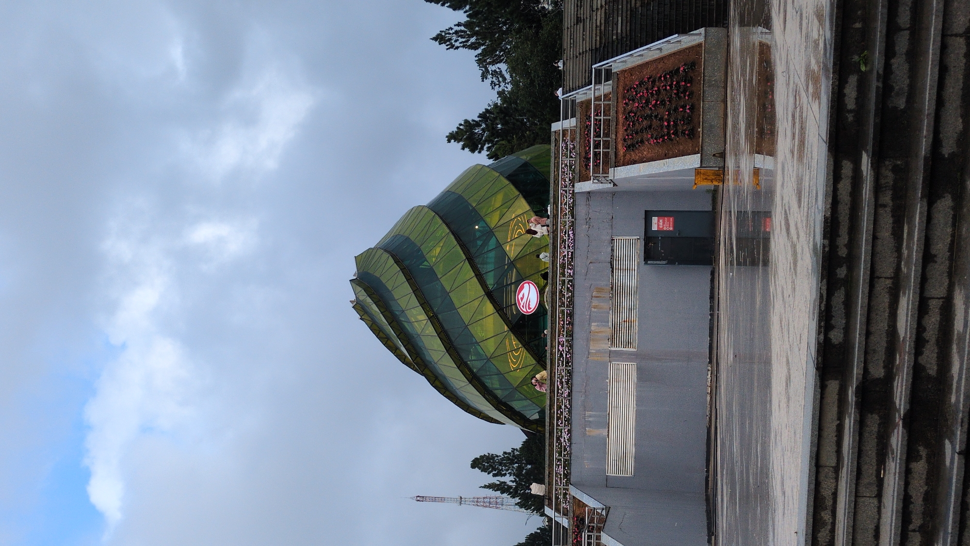 The image shows the exterior of the Da Lat Flower Park's Exhibition House in Da Lat, Vietnam, on a cloudy day. The building, a prominent landmark, has a unique, spiraling green glass facade. In front of the building is a wide, wet plaza with visible reflections on the ground, indicating recent rain. Stone steps lead up to the plaza from the foreground. To the left of the building, a tall, slender radio tower is visible against the overcast sky. Several people are seen standing on a walkway or balcony in front of the building. The scene appears to be a public space, possibly a tourist attraction, with an emphasis on the architectural design of the exhibition house and its surrounding environment.