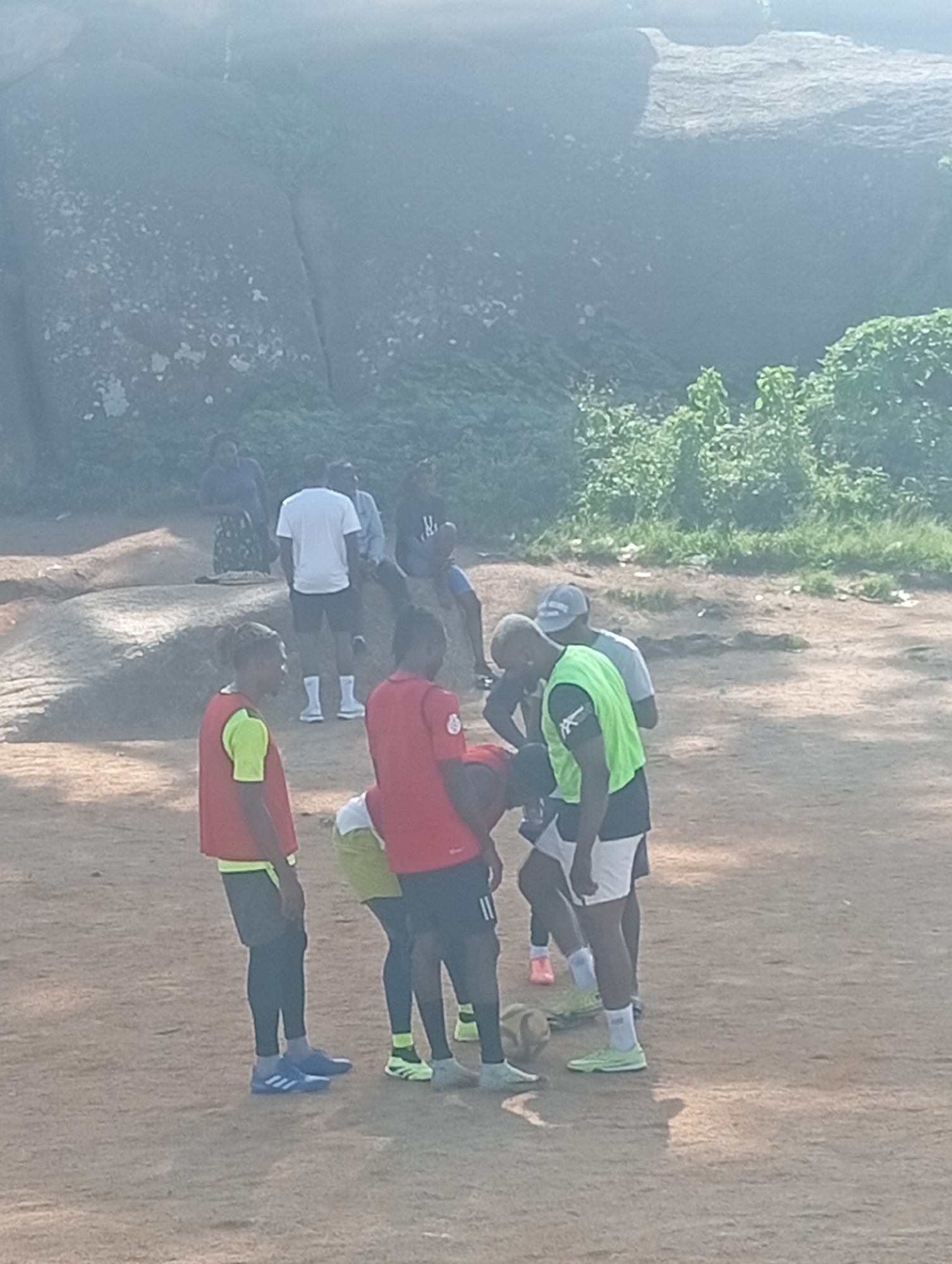 In an outdoor, arid environment, potentially at the Rwang Pam Stadium area in Jos, Nigeria, a group of individuals are gathered on dusty, reddish-brown terrain. Several large, weathered rock formations, some with patches of moss or lichen, dominate the background, interspersed with dense green foliage. Sunlight casts shadows, indicating it is daytime.
In the foreground, five men are engaged in what appears to be a casual soccer practice or discussion. Two men in the center wear bright, contrasting vests: one in neon green, the other in red over a yellow shirt. Both are focused downwards, with the man in the green vest appearing to bend slightly. Another man, partially visible in the foreground right, also wears a red vest. To the left, a man in a red vest and black athletic pants stands looking to his left. In the middle ground, a man in a white t-shirt and black shorts stands with his hands in his pockets, facing away from the camera. Other individuals are present in the background, including a woman in a patterned skirt and dark top, and two men. A soccer ball is visible on the ground near the man in the green vest. The overall atmosphere is informal and active.