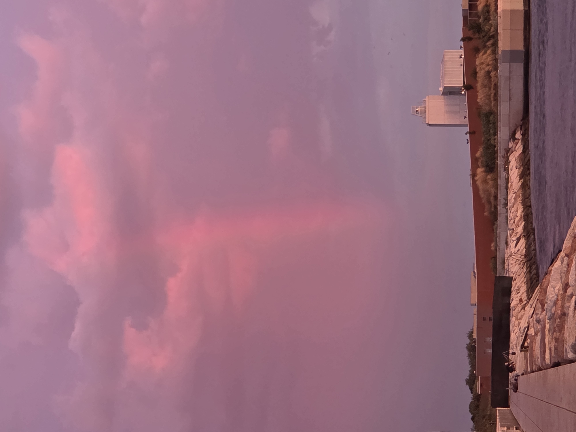 A vertical shot captures a vibrant pink sky over Chioggia, Italy. The sky is filled with soft, cotton-like clouds tinted with hues of magenta and fuchsia, with a distinct vertical shaft of light breaking through the clouds. In the distance, the silhouette of modern buildings, including a tall, rectangular tower, is visible against the horizon. A low, rust-colored wall runs along the foreground, separating the buildings from a rocky embankment. The embankment is made of rough, irregular stones and slopes down to the water. A walkway, possibly a pier or promenade, is visible on the left side of the image, made of light-colored rectangular planks. The water in the foreground is dark and calm, reflecting the colors of the sky. The overall scene evokes a sense of peacefulness and natural beauty, highlighted by the dramatic sunset.