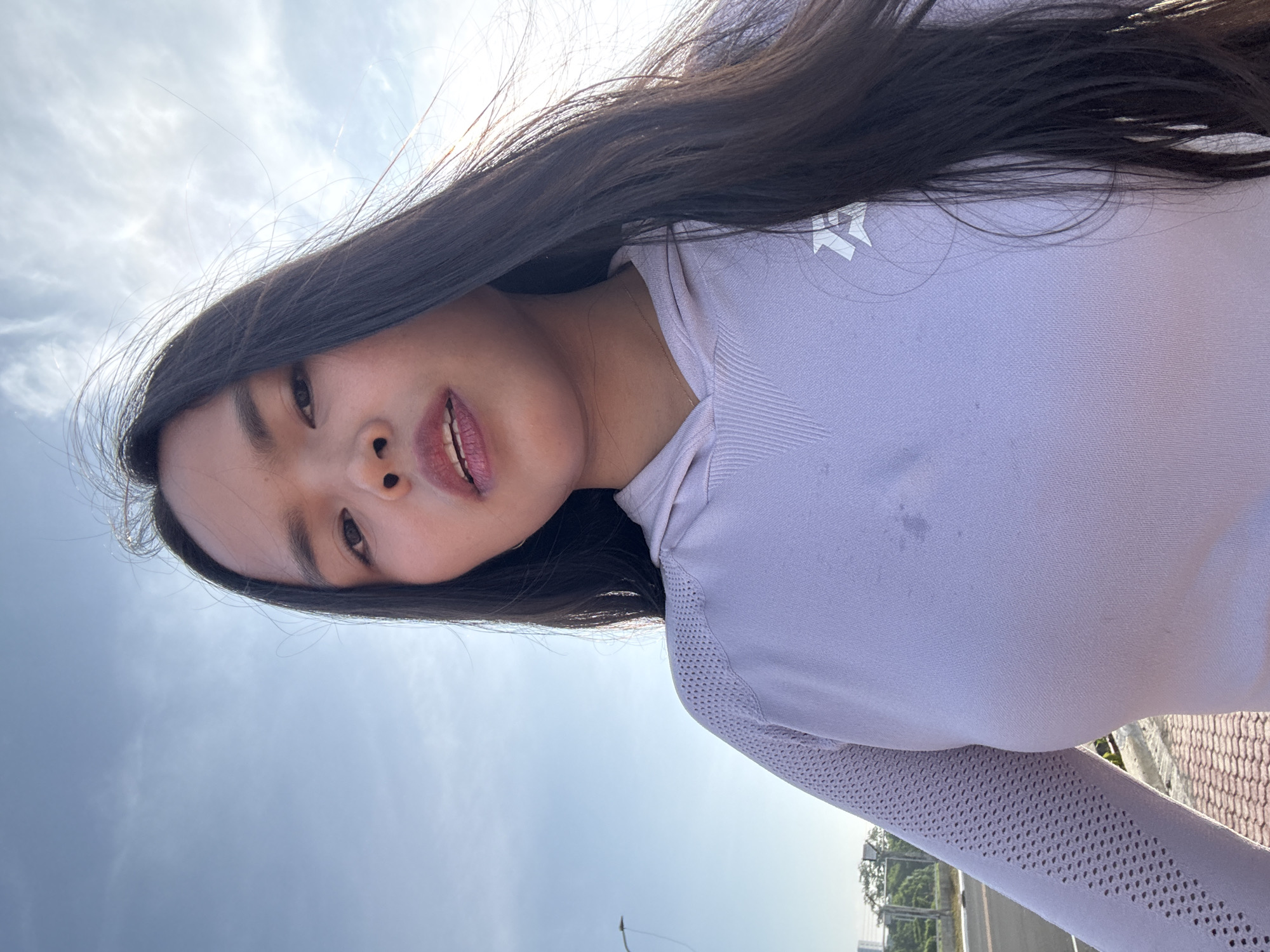 A young woman with long, dark hair and light skin is captured in a close-up, selfie-style shot outdoors against a pale blue sky with scattered clouds. She is wearing a light purple long-sleeved hooded shirt with textured detailing on the sleeves and shoulders, and a white logo on the right chest. Her mouth is slightly open, revealing teeth and red lipstick. Her gaze is directed towards the camera. The background suggests an urban setting, with a paved road, sidewalk, a lamppost, and distant buildings visible. The lighting indicates it is daytime, possibly late afternoon, with the sun casting some glare. The scene is likely located in Davao City, Philippines, given the environmental context and typical urban infrastructure.