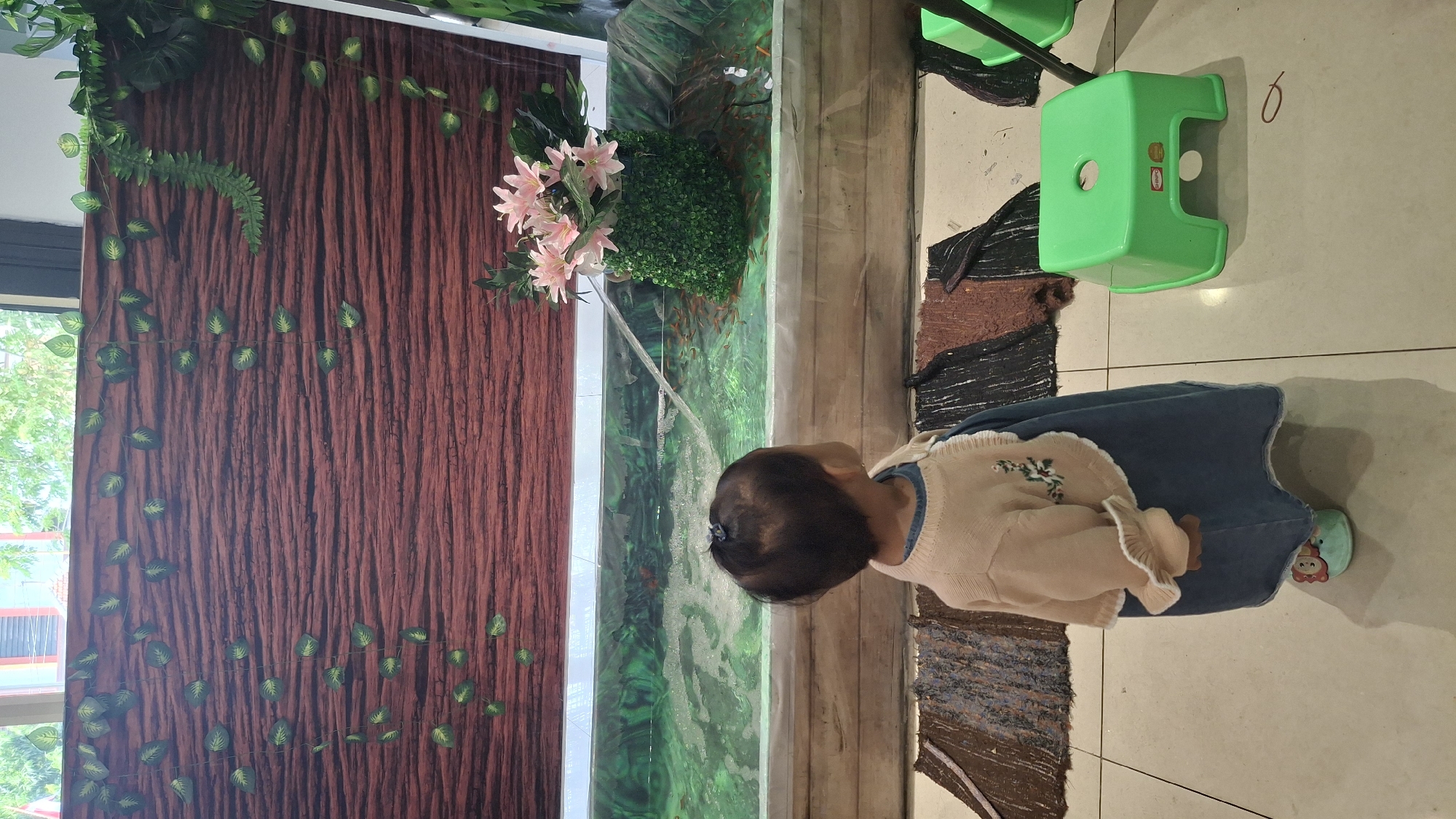 A young child, viewed from behind, stands looking into a large aquarium. The child is wearing a denim dress and a light-colored cardigan with embroidered flowers. The aquarium contains numerous small fish and a decorative arrangement of green artificial foliage topped with pink artificial flowers. A fountain is visible in the aquarium. Behind the aquarium, a backdrop features a wood-grain pattern and artificial green vines. In the foreground, a small green plastic stool is placed on a tiled floor. The scene appears to be an indoor setting, possibly a public space or a home aquarium display, located in Kecamatan Pati, Indonesia.