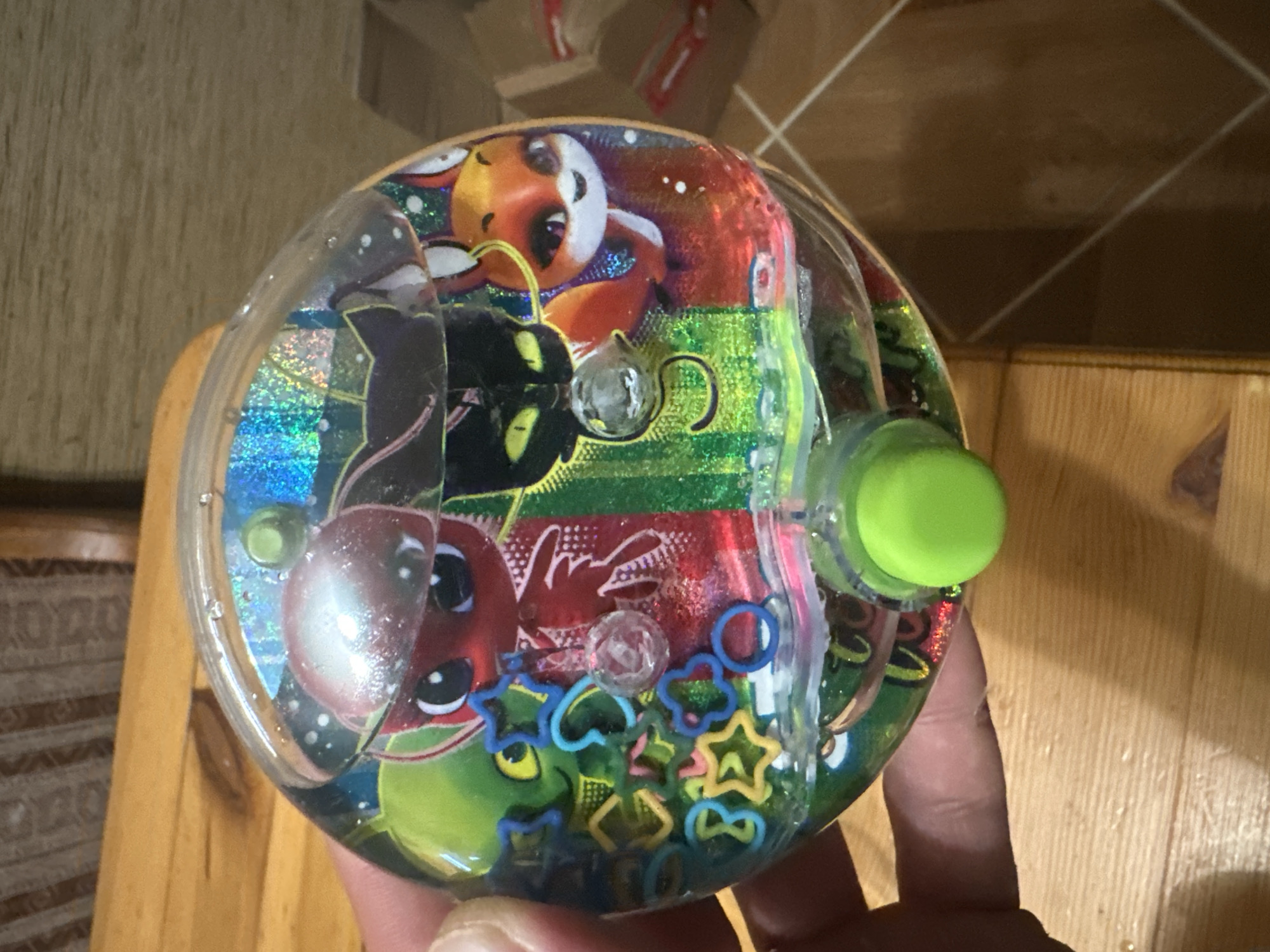 A person's hand holds a clear, spherical plastic toy filled with colored liquid and small, decorative plastic pieces. The toy features images of cartoon characters, including a red character with a silver helmet, a black cat with green eyes, and an orange fox with white accents. The characters are superimposed over a background with shimmering blue and green stripes. Inside the toy, small, colorful plastic shapes such as stars, circles, and hearts are suspended in the liquid, along with some loose blue rings. A green button is visible on the lower portion of the toy. The background suggests an indoor setting with wooden furniture. The location is not depicted in the image.