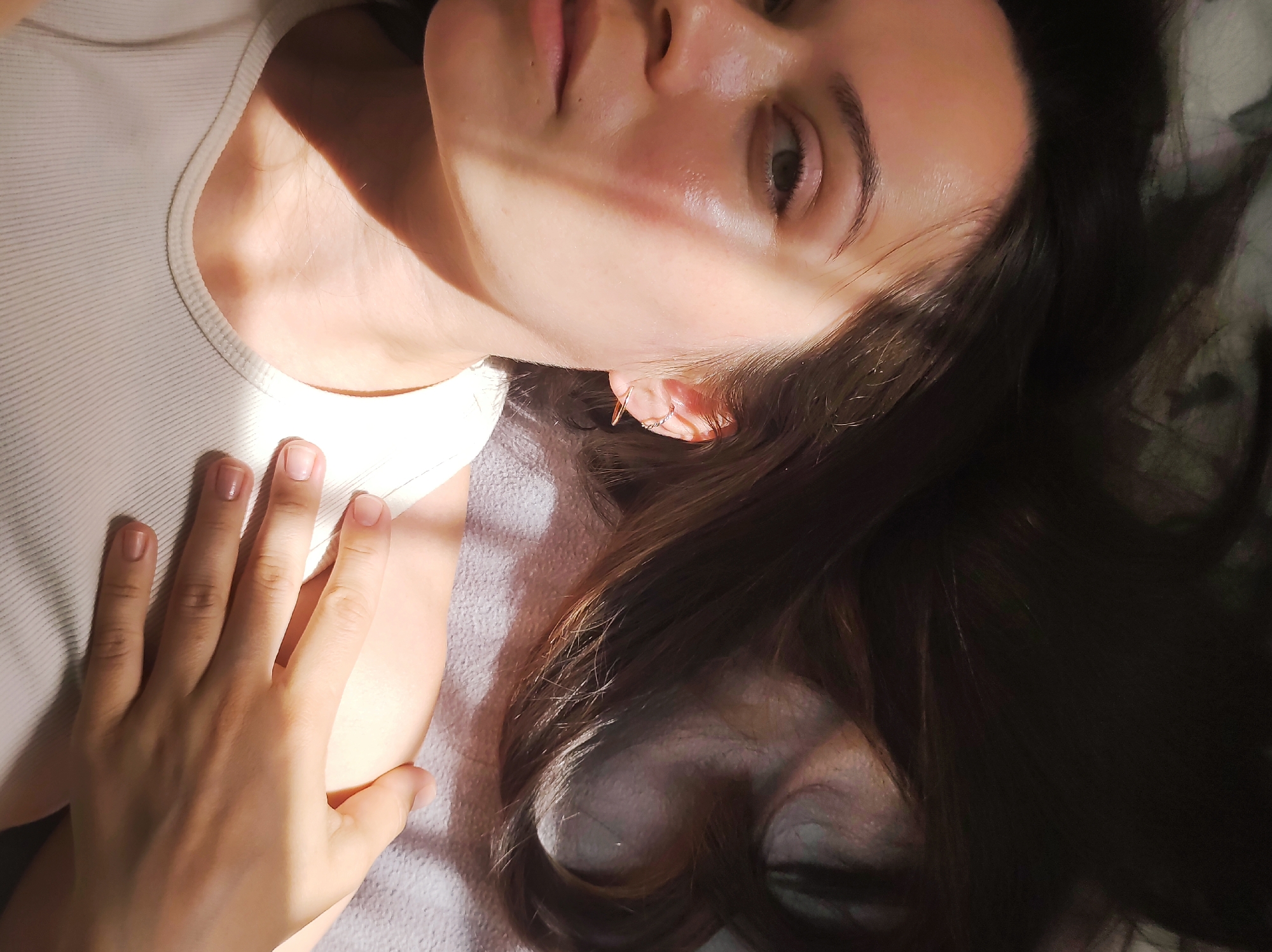A woman is lying down, facing the camera at a slight angle. Her dark hair is spread out around her head. She has fair skin, and her right hand rests on her chest. She is wearing a white tank top. Light and shadow fall across her face. The background appears to be a grey surface, possibly a bed or blanket. It is not possible to identify the location as Chaikovsky, Russia based on the image.