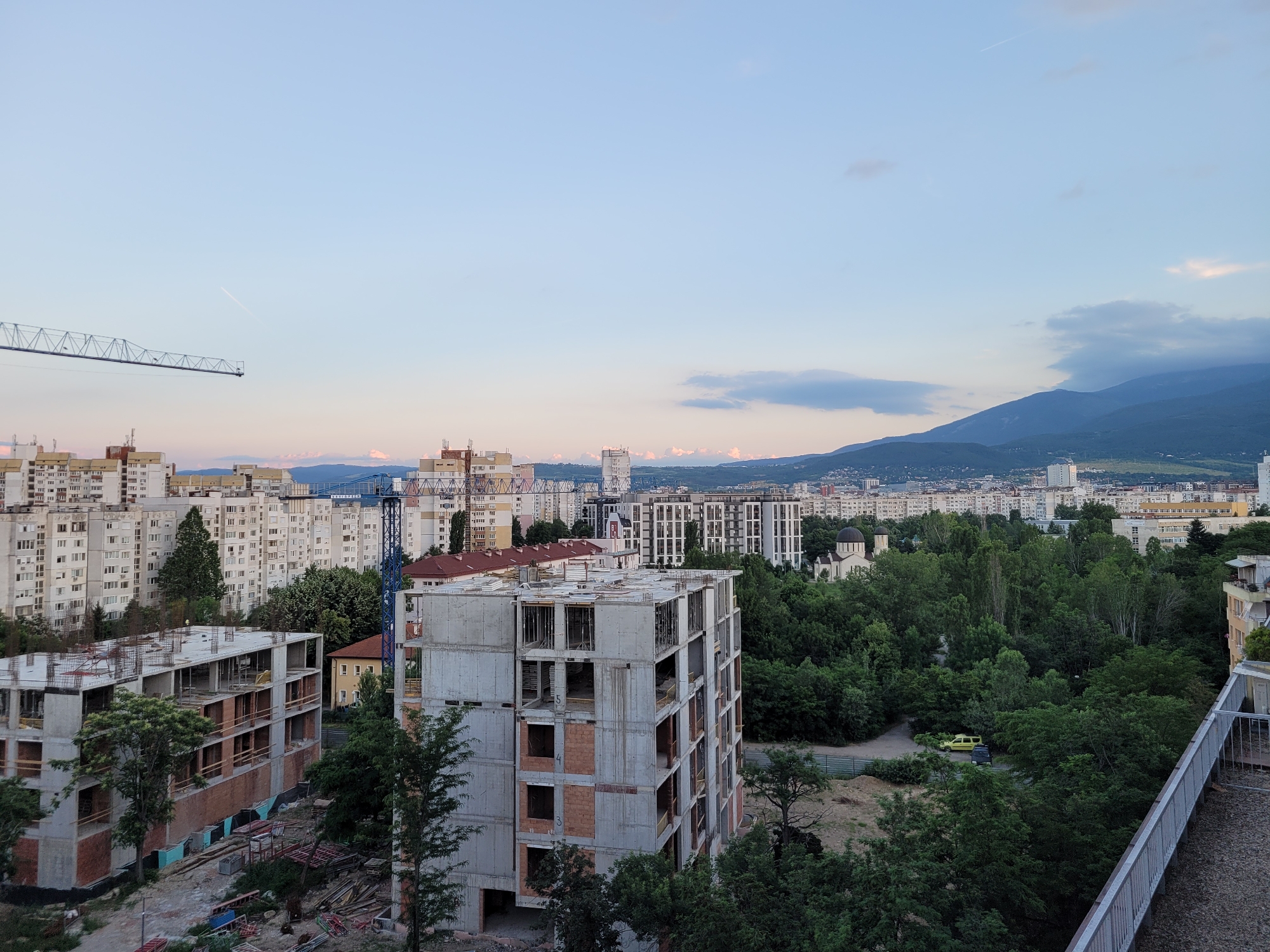 The image depicts an urban landscape in Sofia, Bulgaria, as seen from a high vantage point. The foreground features a multi-story building under construction, characterized by exposed concrete and brickwork. Several other buildings, some completed and others still in various stages of construction, are visible throughout the scene, including apartment blocks and larger modern structures. A prominent blue construction crane is situated to the left of the frame, extending towards the sky. Beyond the buildings, a dense expanse of lush green trees forms a significant natural element within the cityscape. In the distance, a range of mountains forms the horizon line, with a clear sky overhead, suggesting it is either early morning or late evening, indicated by the soft lighting and pastel hues in the sky. A thin white contrail from an aircraft is visible high in the sky. Two cars, a yellow and a dark vehicle, are parked on a road amidst the greenery. A small church with a dome is also visible within the treeline.