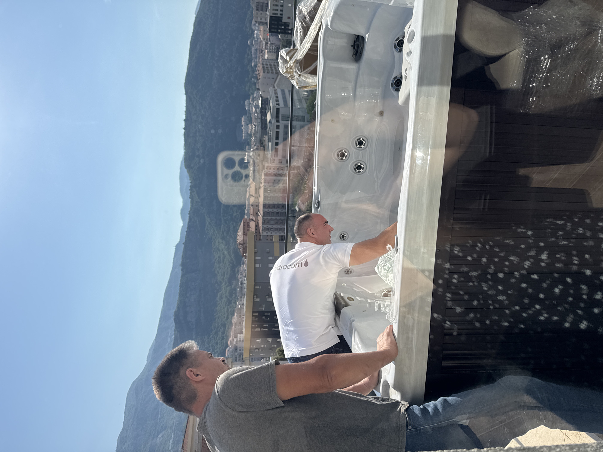 Two men are installing a new hot tub on a high-rise balcony in Budva, Montenegro, with a panoramic view of the city and surrounding mountains. One man is wearing a gray t-shirt and blue jeans, while the other is wearing a white t-shirt with the text "Hydrocom" on the back and dark shorts. The hot tub is wrapped in plastic, but the jets are visible. The sun is shining brightly. A smartphone is visible, reflecting on the surface of the glass balcony fence.