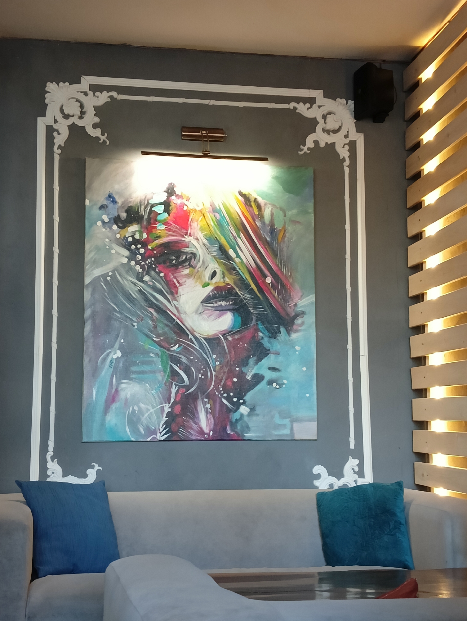The image shows a dimly lit interior space, likely a cafe or lounge, in Saint Petersburg, Russia. A grey wall serves as the backdrop, adorned with an abstract painting of a woman's face in vibrant, psychedelic colors. The painting is framed by white decorative molding. Above the painting, a modern brass light fixture illuminates the artwork. To the right of the painting, a speaker is mounted on the wall, and a slatted wooden panel with integrated lighting adds a warm, ambient glow to the scene. In the foreground, a light grey sofa is visible, with two decorative cushions, one blue and one teal. The overall atmosphere is contemporary and artistic.