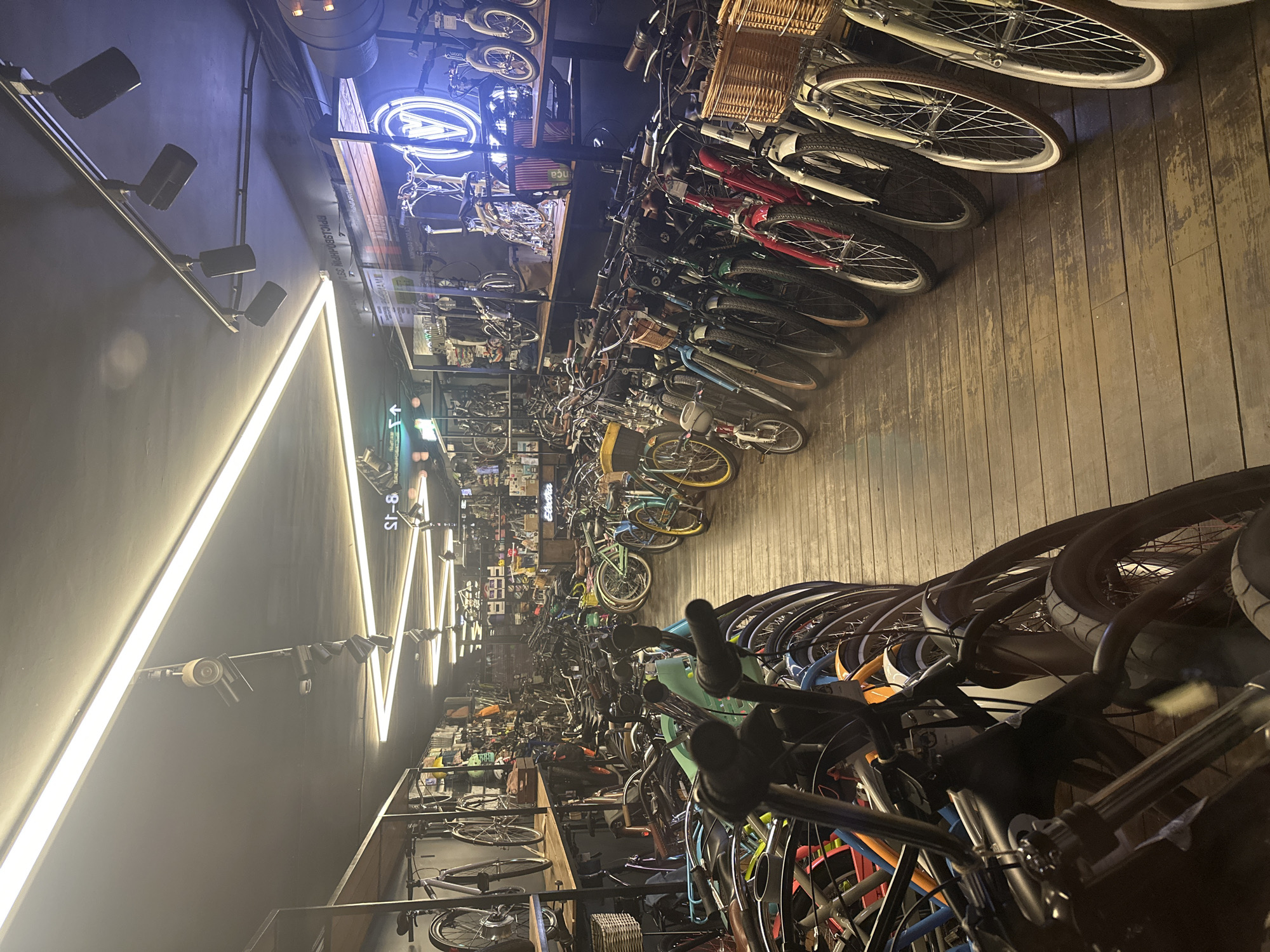 The image shows the interior of a bicycle shop. Rows of bicycles are arranged along the top and bottom edges of the frame, obscuring the center of the image. The shop has a wooden floor. On the left, a black wall is adorned with triangle-shaped neon lighting fixtures and track lighting. At the top of the image, a neon sign featuring the letter "A" is visible. The image appears to have been taken in Moscow, Russia, as indicated by the Russian text on a sign in the background.