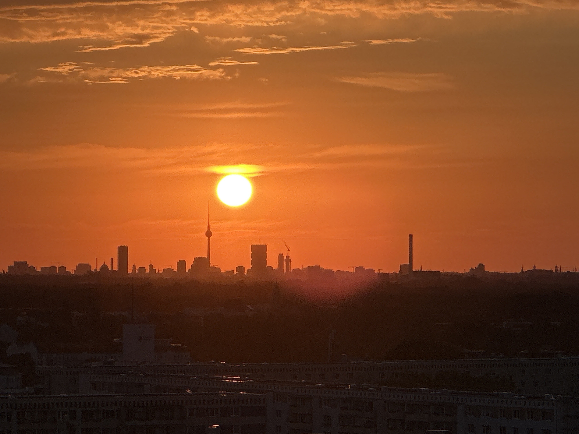 This is a photograph of the Berlin skyline at sunset. The sun, a bright white orb, is positioned just above the horizon, casting a warm orange glow across the sky. Wispy clouds are visible in the upper part of the frame, also tinged with orange. In the mid-ground, the silhouettes of various buildings form the city's skyline. Most prominent are the Fernsehturm (TV Tower) and a tall, rectangular skyscraper. Several other buildings of varying heights are also visible, some with visible cranes, suggesting ongoing construction. In the foreground, the dark shapes of apartment buildings are visible, their details obscured by the low light. The overall impression is of a city bathed in the dramatic light of a setting sun.