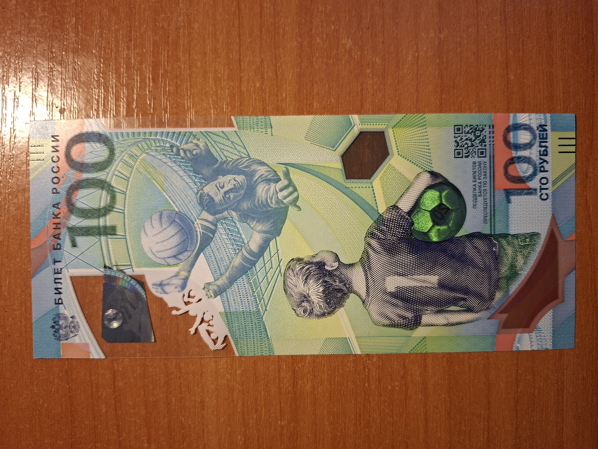 The image shows a close-up, top-down view of a Russian 100 ruble banknote with a football (soccer) theme, placed on a wooden surface. The banknote features a stylized depiction of a boy looking up at a football player in mid-air, both rendered in a graphic, line-art style. A football is prominently displayed on the banknote, with one held by the player and another near the boy. The banknote has holographic security features, including a small soccer ball within a circular holographic patch. Text on the banknote is in Russian, with "БИЛЕТ БАНКА РОССИИ" (Bank of Russia Ticket) and "100" being the most prominent. Below the image, it reads "СТО РУБЛЕЙ" (One Hundred Rubles). The wooden surface has visible grain patterns and a slightly reflective quality, suggesting it might be a table or desk. The lighting appears to be from above, casting subtle shadows.