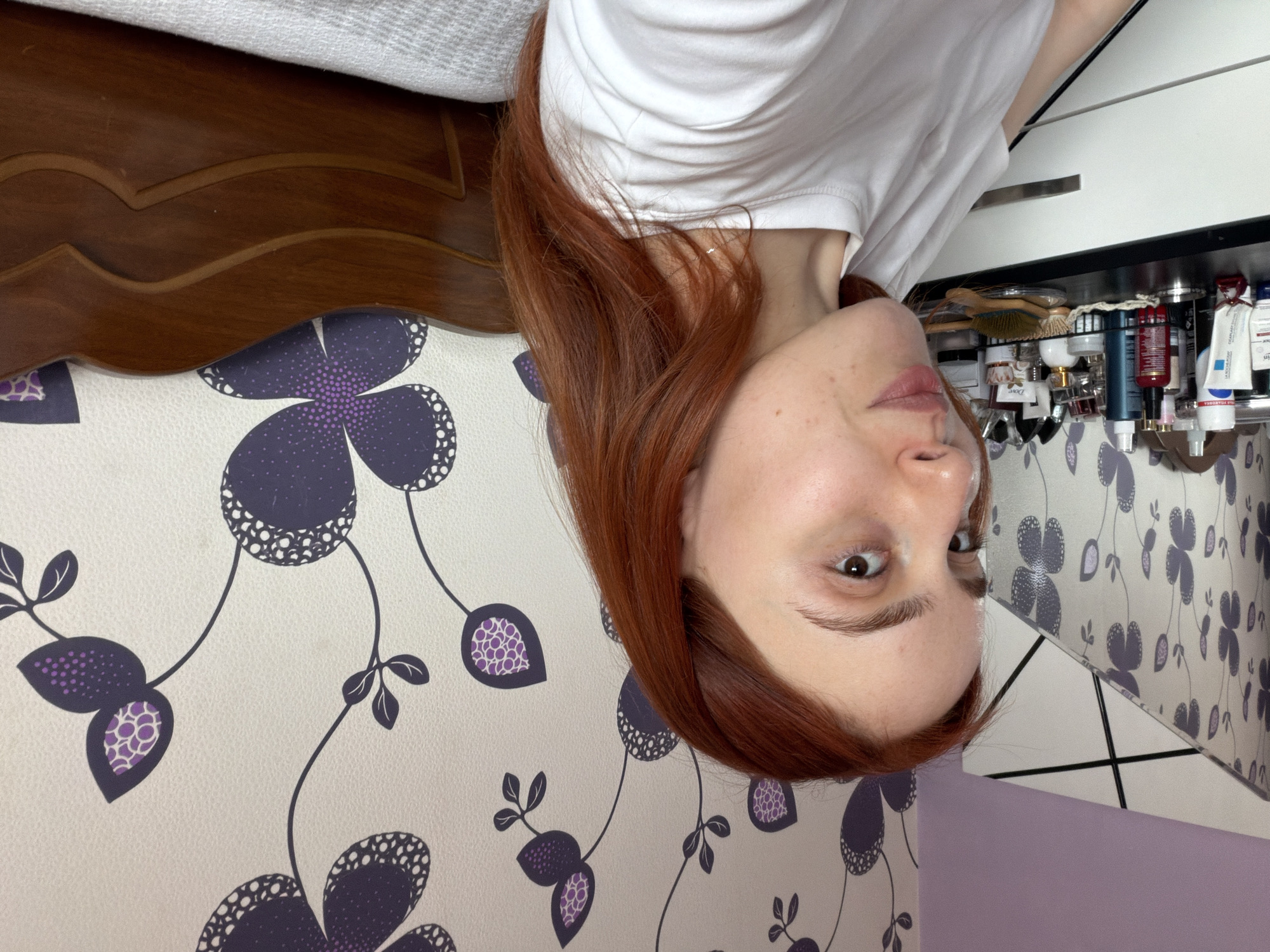 The image shows a woman with auburn hair, wearing a white T-shirt, lying on a bed with a patterned headboard and floral wallpaper. Adjacent to the bed is a white dresser with various toiletries and cosmetic products displayed on the shelf above. The location cannot be determined from the image.