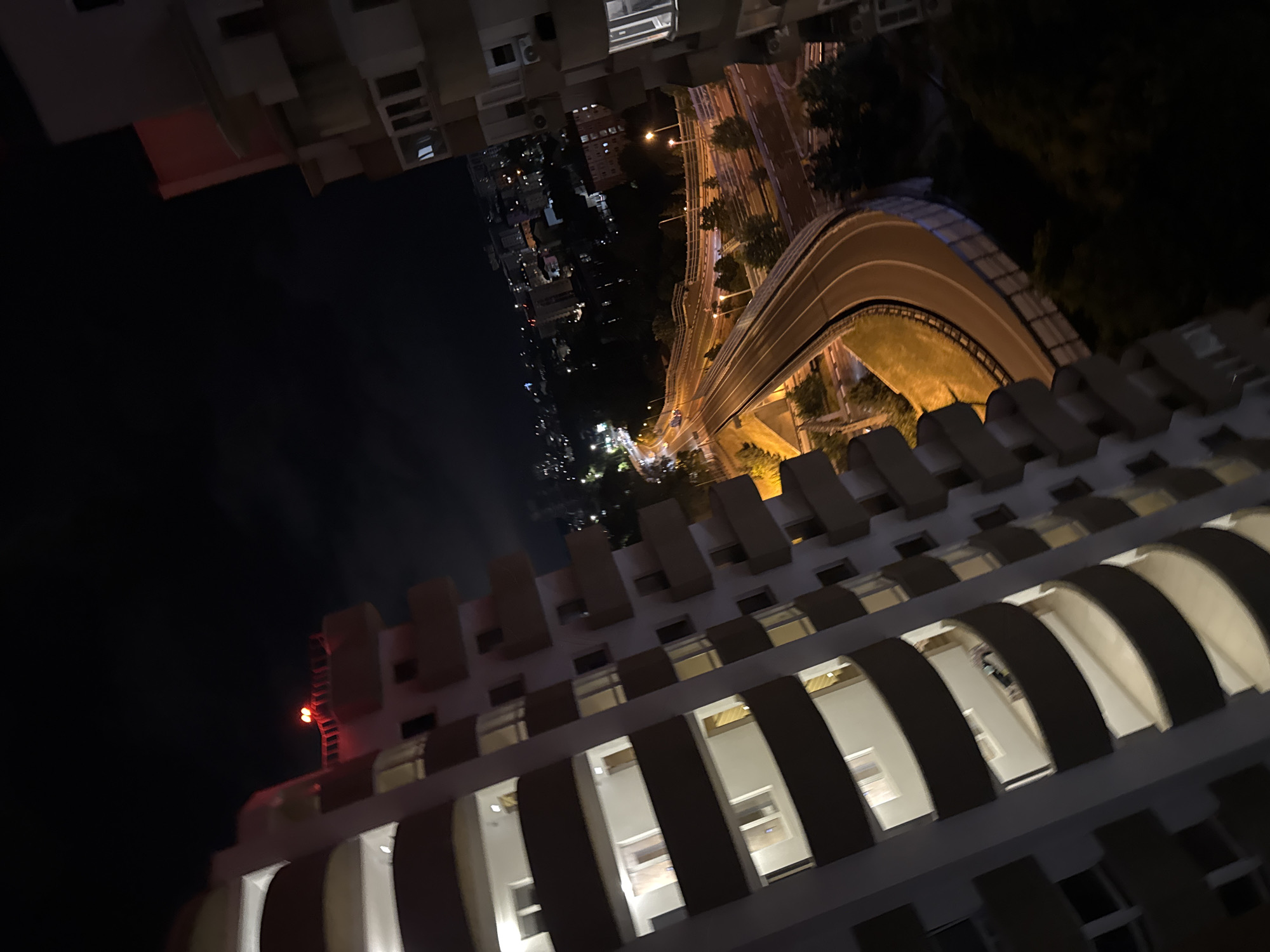 The image depicts a nighttime aerial view of a city, likely in Sochi, Russia, with a focus on a multi-lane highway curving through the urban landscape. The foreground is dominated by the side of a tall apartment building with curved balconies, its windows illuminated. Two other apartment buildings are visible, one to the right and another in the distance. Red lights are visible atop one of the buildings, possibly for aviation warning. The highway is well-lit by streetlights, and cars are present on the road. Trees and other urban structures are scattered throughout the scene. The overall impression is of a bustling city at night.