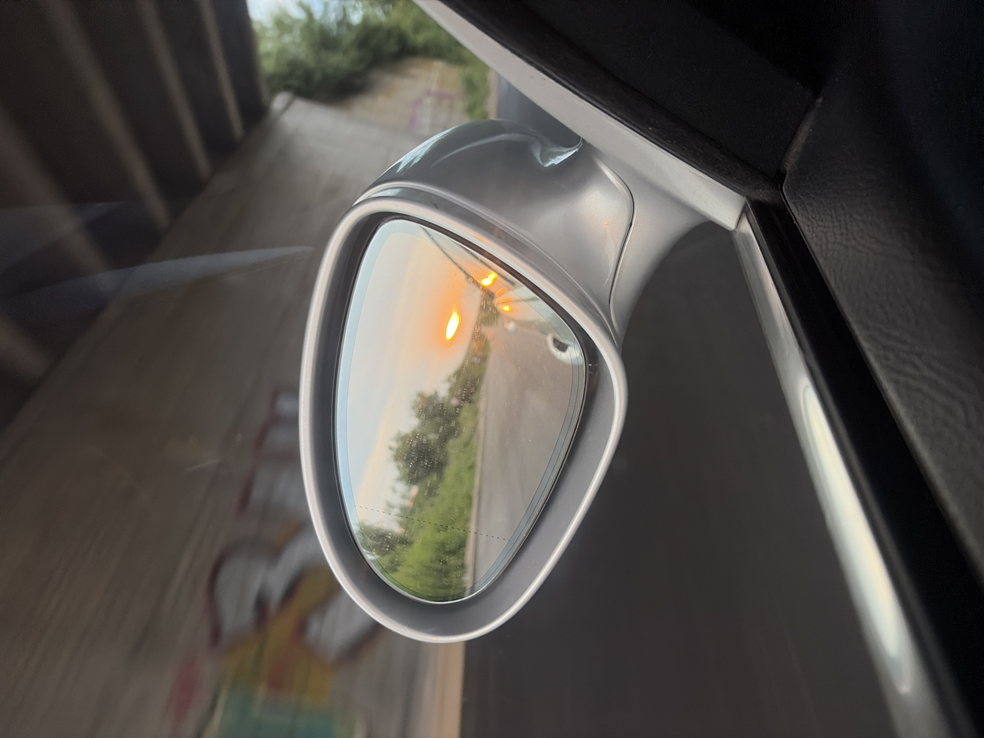 This image captures a rear-view mirror reflection of a sunset, viewed from inside a moving vehicle. The vehicle appears to be under an overpass with graffiti visible on the concrete structure above. The reflection shows a road with greenery on the side, and the setting sun casting an orange glow, with several points of light indicating oncoming vehicle headlights. The foreground of the image shows the interior of the car, including the door panel and the side mirror assembly. The location is identified as Vasil Levski, Bulgaria.