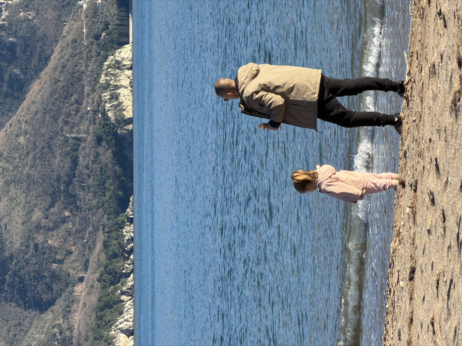The image captures a serene outdoor scene featuring two individuals on a pebbly beach next to a large body of water, with forested mountains in the background under bright daylight.
In the foreground, a man and a young child are positioned near the water's edge. The man stands taller, wearing a light brown or khaki long-sleeved jacket, dark pants, and dark sneakers with white soles. He has short hair or is bald and is looking down towards the water. His right hand appears to be holding a light-colored object, possibly a piece of food or a stone, which he might be offering or dropping into the water.
Slightly behind and to the left of the man, a small child stands closer to the water. The child is dressed in a light pink hooded jacket and matching pants, with light-colored shoes. The child is also looking down intently at the water, near where a small wave is receding from the shore. Both individuals seem engaged in observing or interacting with the water.
The body of water, likely a sea or a large lake, is a vibrant blue-green color with visible ripples and small, gentle waves breaking along the shoreline. The water appears calm and reflective under the sunny conditions. The beach itself is composed of light brown and tan pebbles and possibly some coarse sand, stretching from the water's edge into the foreground.
In the background, majestic mountains rise steeply from the opposite shore of the water. These mountains are densely covered with dark green vegetation, primarily trees, and feature some visible rocky sections. Hints of structures or paths can be faintly seen on the mountain slopes, suggesting human presence or development in the distance.
The lighting indicates a clear, sunny day, likely during late morning or early afternoon, with strong shadows and bright illumination across the scene. The attire of the man and child suggests it might be a cool but pleasant day for outdoor activities by the water. There is no visible text in the image.