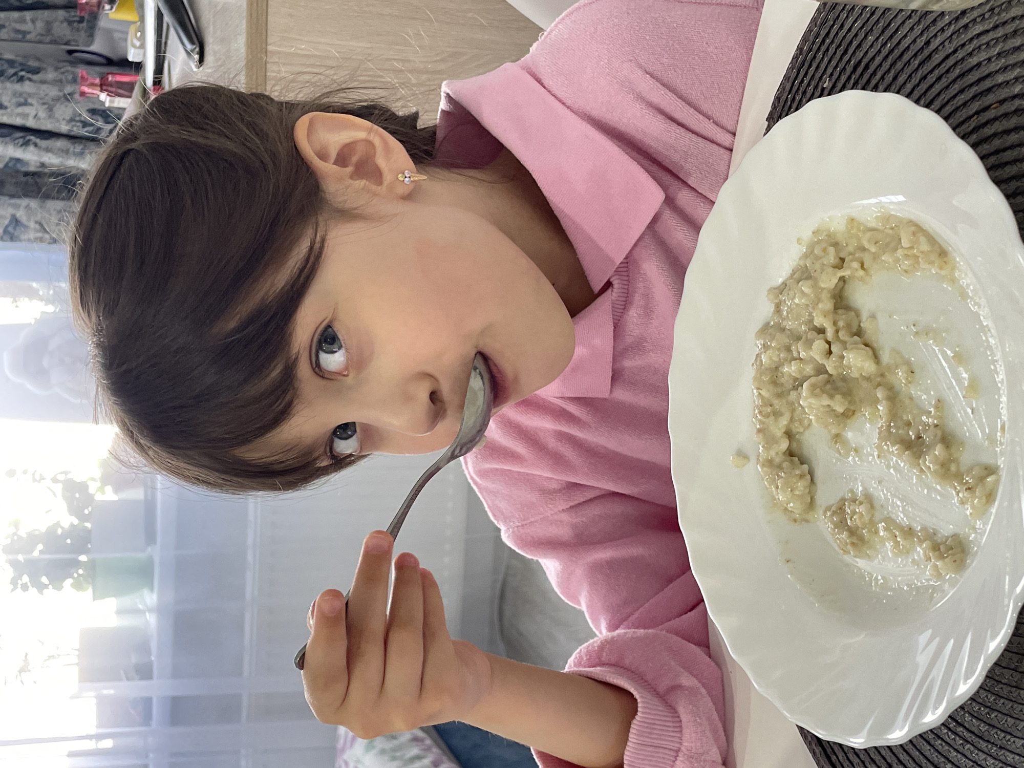 A young girl wearing a pink sweatshirt is eating a bowl of oatmeal. The oatmeal is in a white plate with a scalloped edge. The girl is holding a metal spoon in her right hand, bringing it to her mouth. She has brown hair and blue eyes. Her right ear is visible, with a small earring. The plate is sitting on a dark gray, woven placemat. In the background, there is a window with sheer white curtains. Based on the location provided, this scene is likely taking place indoors in Izhevsk, Russia.