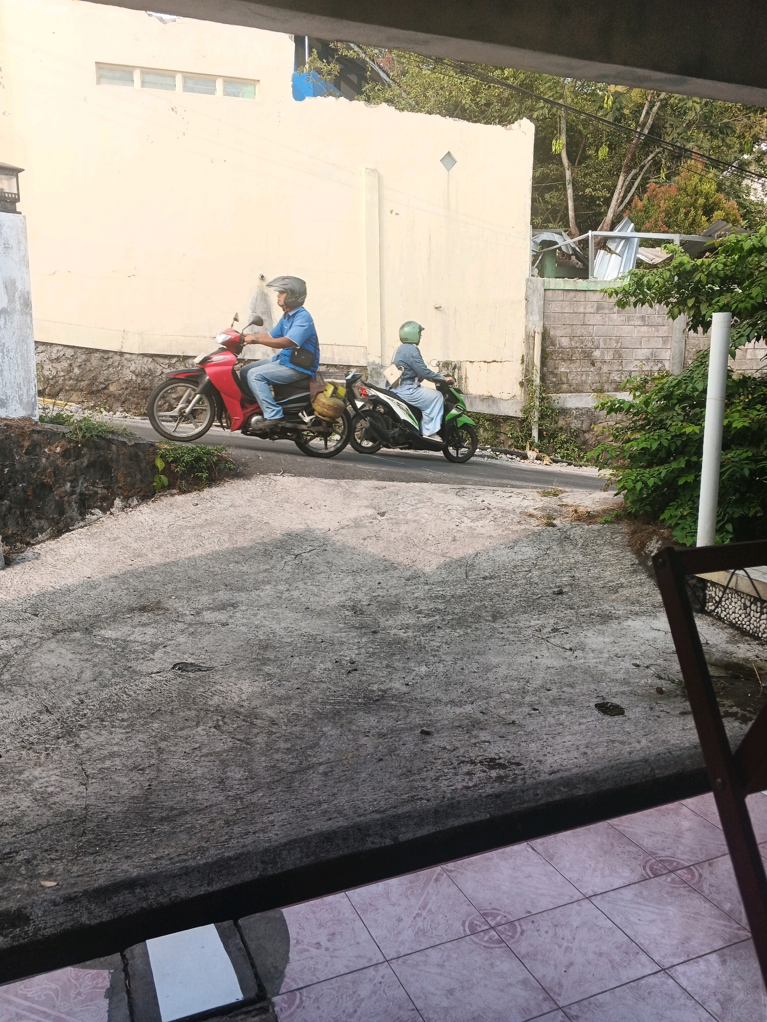 Two individuals on motorcycles are captured from a slightly elevated vantage point, possibly a balcony or porch, looking down onto a sloping concrete path. The scene is set in Keji, Indonesia, characterized by its residential architecture and lush tropical vegetation.
The foremost motorcycle, a vibrant red, is ridden by a person wearing a blue shirt and jeans, their head protected by a dark helmet. A dark bag is slung across their torso, and a yellow bag is secured to the rear of the motorcycle. The second motorcycle, predominantly green, is occupied by an individual in a light blue outfit and a green helmet. A light-colored bag is visible on their shoulder. Both riders appear to be moving away from the viewer, towards the right of the frame.
The environment features a light-colored building with a textured wall, marked by what appears to be a small, square vent. The ground is paved with a rough concrete surface, showing signs of wear and tear. A low stone wall runs alongside the path, bordered by patches of grass and small plants. In the background, a brick wall and dense green foliage indicate a typical urban landscape in a tropical region. Overhead, trees with visible branches and leaves, some bearing fruit or seed pods, suggest the presence of mature vegetation. A partially constructed structure with corrugated metal roofing is also visible behind the brick wall, hinting at ongoing development or repairs. The overall lighting suggests it is daytime, with the sun casting shadows across the concrete. The foreground reveals patterned floor tiles and the edge of a dark-colored structure, possibly furniture, implying the viewpoint is from a sheltered area overlooking the outdoor scene.