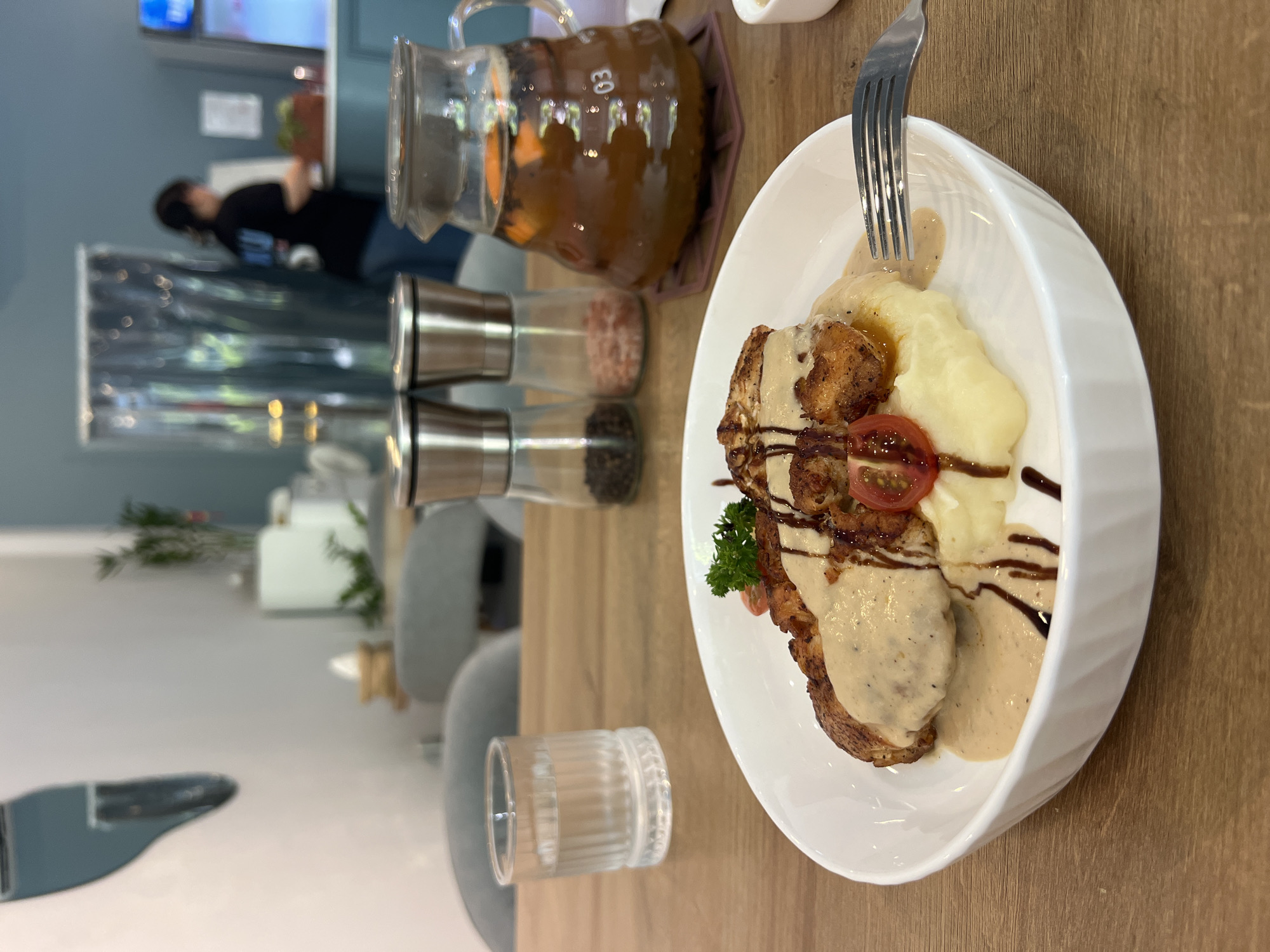 A plate of food with mashed potatoes and fried chicken covered in gravy, a fork sticking into the potatoes, a jar of iced tea with fruit inside, three spice shakers, and a small glass of water are on a wooden table. In the background are chairs and a person, possibly in a cafe or restaurant. Location: Unknown, Unknown.
