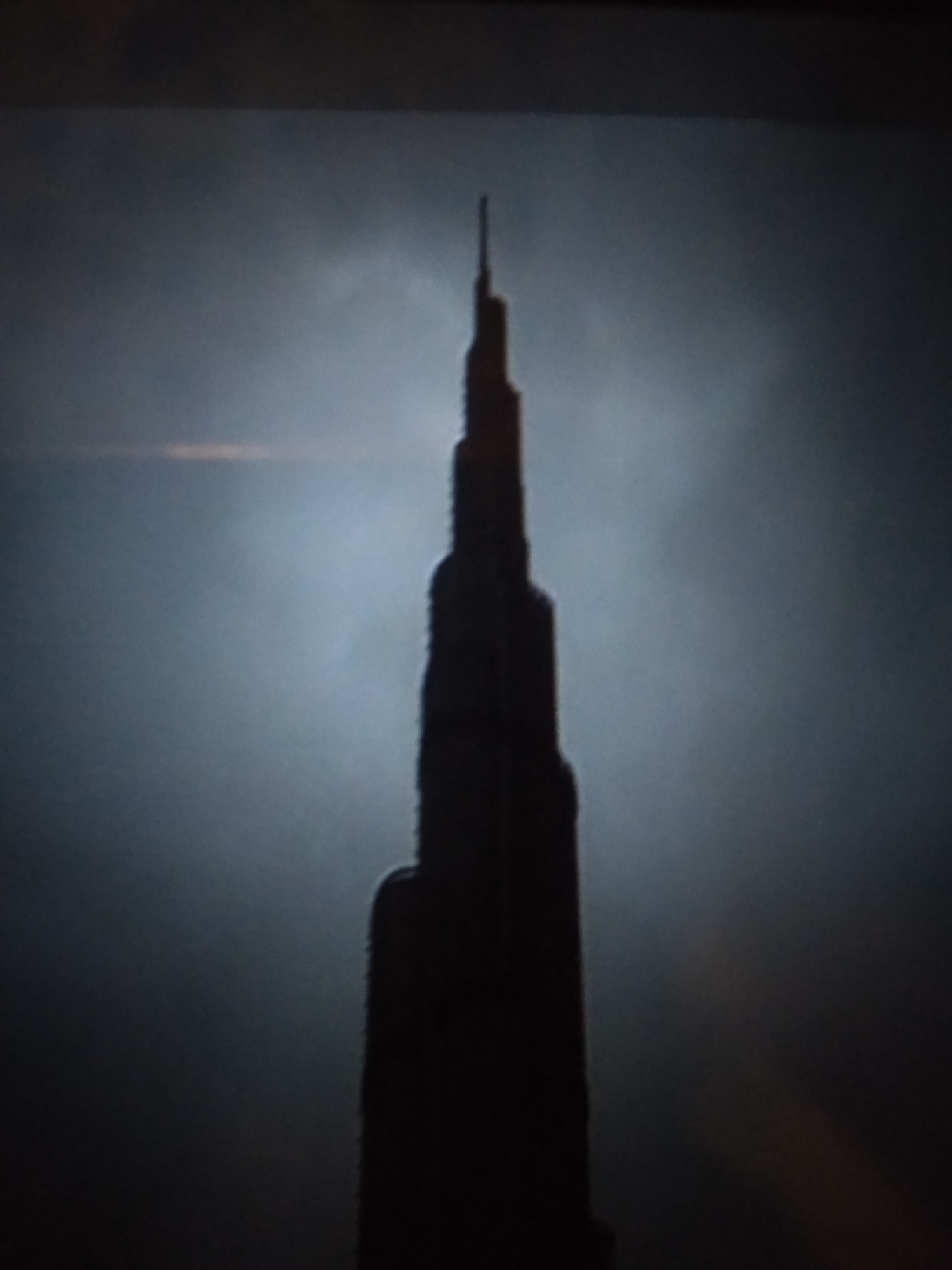 The media file is a photograph of a very tall skyscraper, silhouetted against a hazy, possibly cloudy or foggy sky. The building is dark and its shape suggests a modern architectural design with multiple setbacks, tapering towards a spire at the very top. The lighting is dim, with the bright sky behind creating a strong contrast. There appears to be a faint horizontal band of light across the sky, potentially indicating a sunrise or sunset, or perhaps artificial lighting within the haze. The overall mood is somewhat dramatic and imposing due to the silhouette and the atmospheric conditions. The image is grainy, suggesting it may have been taken in low light or with a low-resolution camera. There are no people or other objects clearly visible in the frame. The setting appears to be urban, given the presence of a skyscraper, but specific location cues are limited to the dark silhouette against the sky. The time of day is ambiguous but leans towards dawn or dusk. No text is visible in the image.