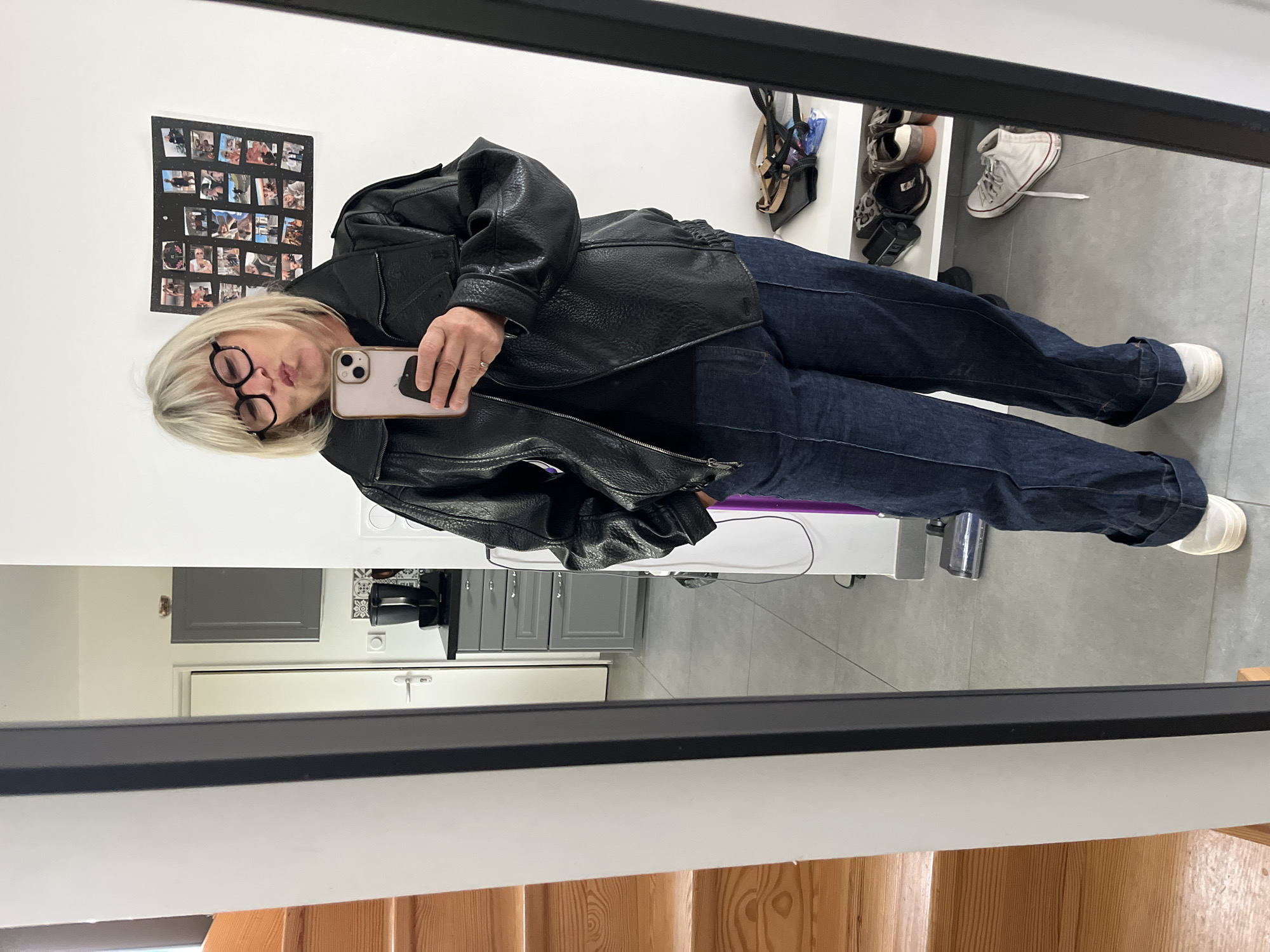 A woman is taking a selfie in a mirror. She is wearing a black leather jacket, dark blue jeans, and white sneakers. She has short blonde hair and glasses, and is making a kissing face. The background includes a wall with pictures, a shoe rack, a kitchen with gray cabinets, and a staircase with wooden railings. The location is in Chambœuf, France.