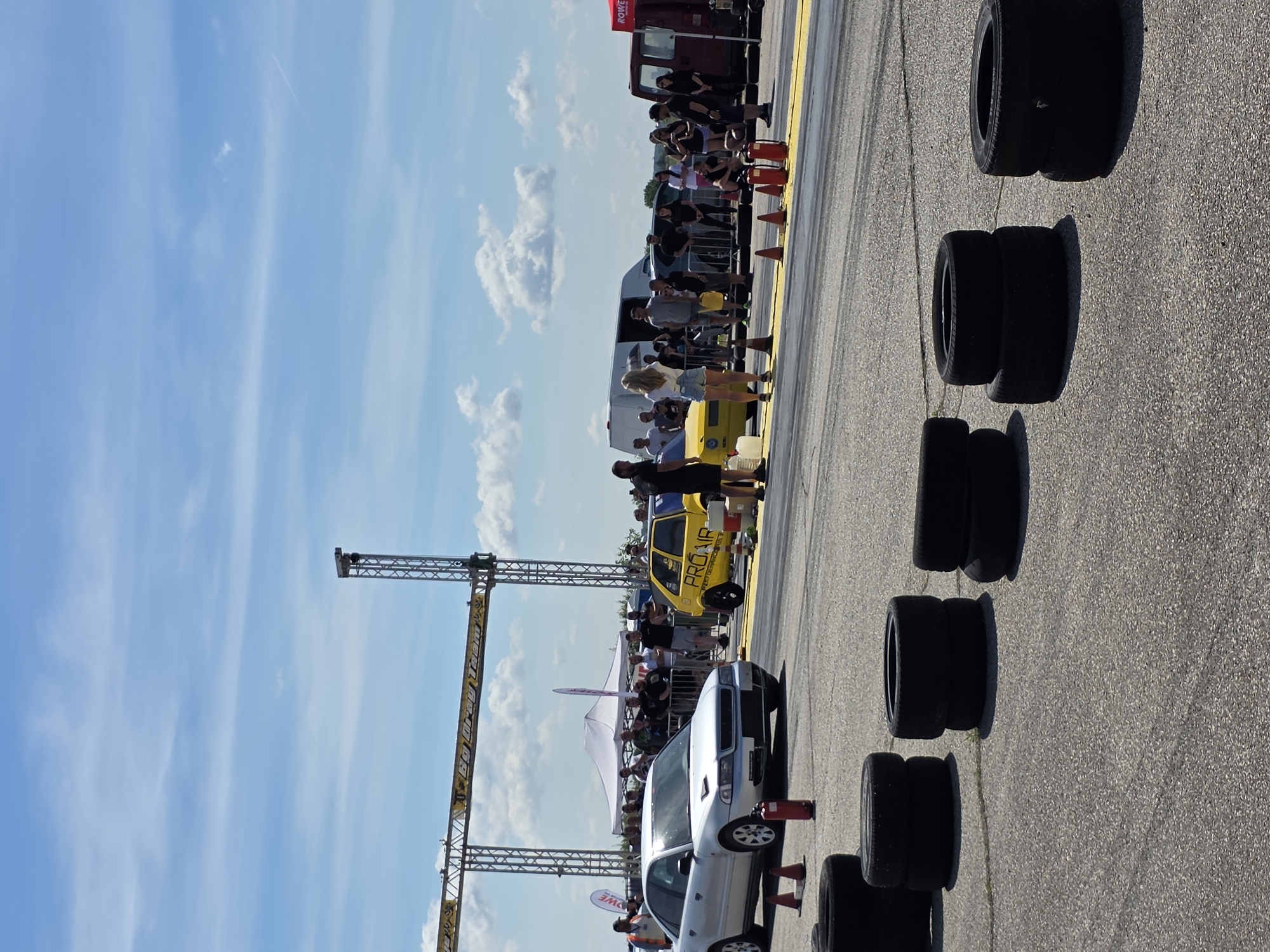 The image depicts a drag racing event on a sunny day, with a bright blue sky and scattered clouds. The scene is set on a paved track marked with yellow lines, characteristic of a drag strip. In the foreground, stacks of tires are placed on the asphalt. Further down the track, a silver sedan and a yellow hatchback are positioned at the starting line. Spectators are gathered behind a barrier, observing the event. Tents and trailers are visible in the background, suggesting a functional setup for a racing event. The overall environment appears to be an open area, possibly an airfield or a dedicated racing facility. Based on the context of the event, it is highly probable that this drag racing event is taking place in Akatsievo, Bulgaria, which is known for hosting such competitions.
