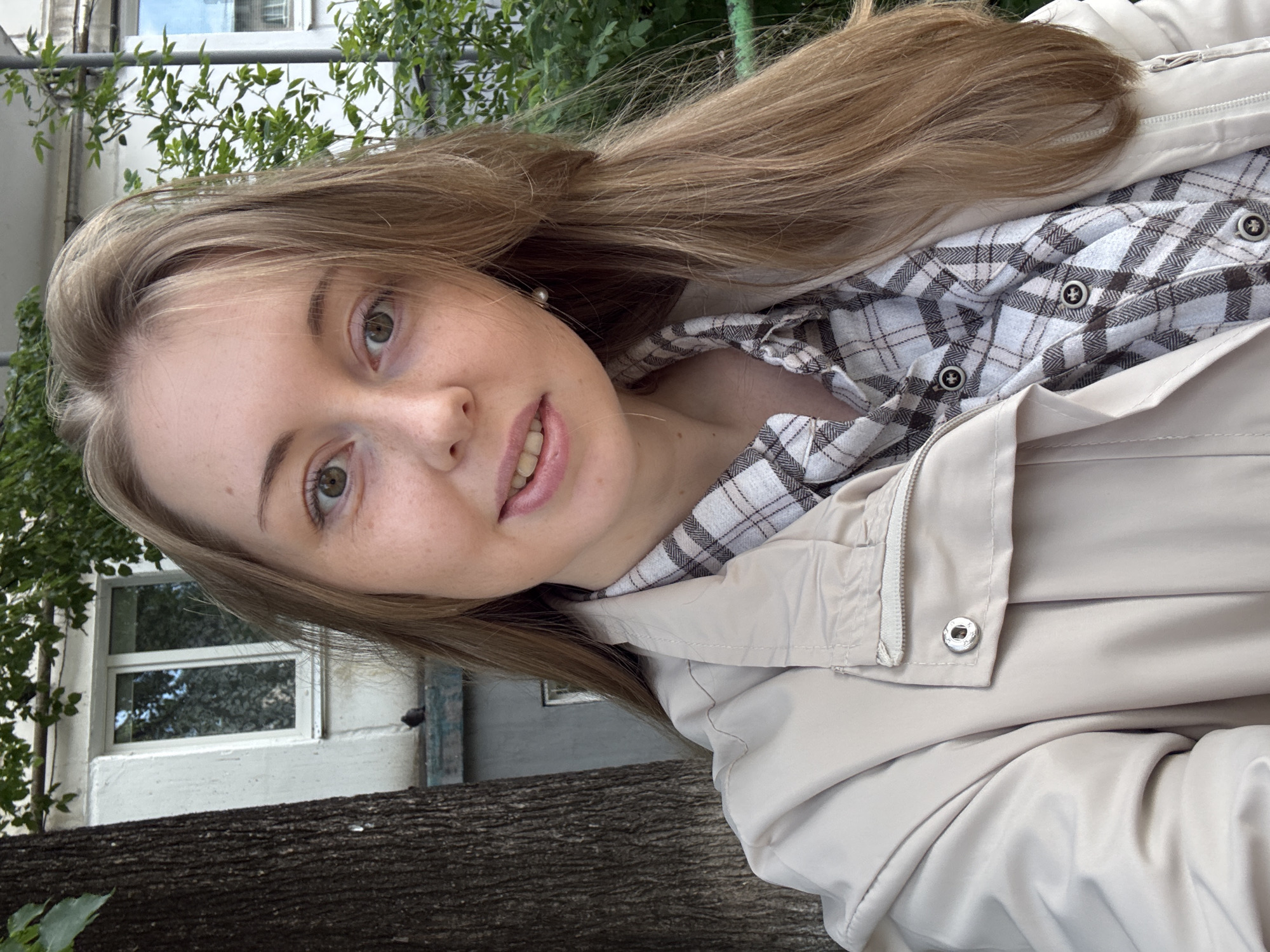 A close-up, slightly rotated, self-portrait features a young woman with fair skin and long, light brown hair, standing outdoors in Unknown, Unknown. She is wearing a light beige jacket over a gray and white plaid shirt, only partially visible. Her face is centered in the frame, revealing light eyebrows, green eyes, and subtle freckles. A hint of pink lipstick is visible on her lips.
The background includes green foliage and glimpses of a light-colored building with a window, partially obscured by the tree. A rough, dark, wooden surface is visible in the lower part of the frame. The overall lighting appears soft and natural.