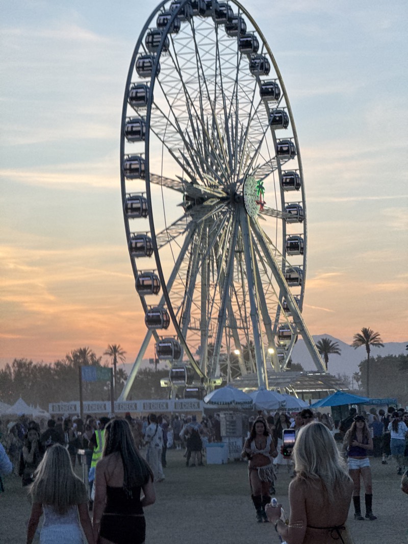 The image depicts a scene at the Coachella Valley Music and Arts Festival in Indio, California. A large Ferris wheel dominates the background, with numerous passenger cars visible. The sky transitions from orange near the horizon to a lighter blue above. In the foreground, a crowd of people is gathered, with several individuals more prominently featured. Two women in the foreground are walking away from the camera, while another woman is taking a photo with her phone. Several tents or canopies are visible in the middle ground. Palm trees are scattered throughout the scene and in the distant landscape. The lighting suggests it is either sunrise or sunset.