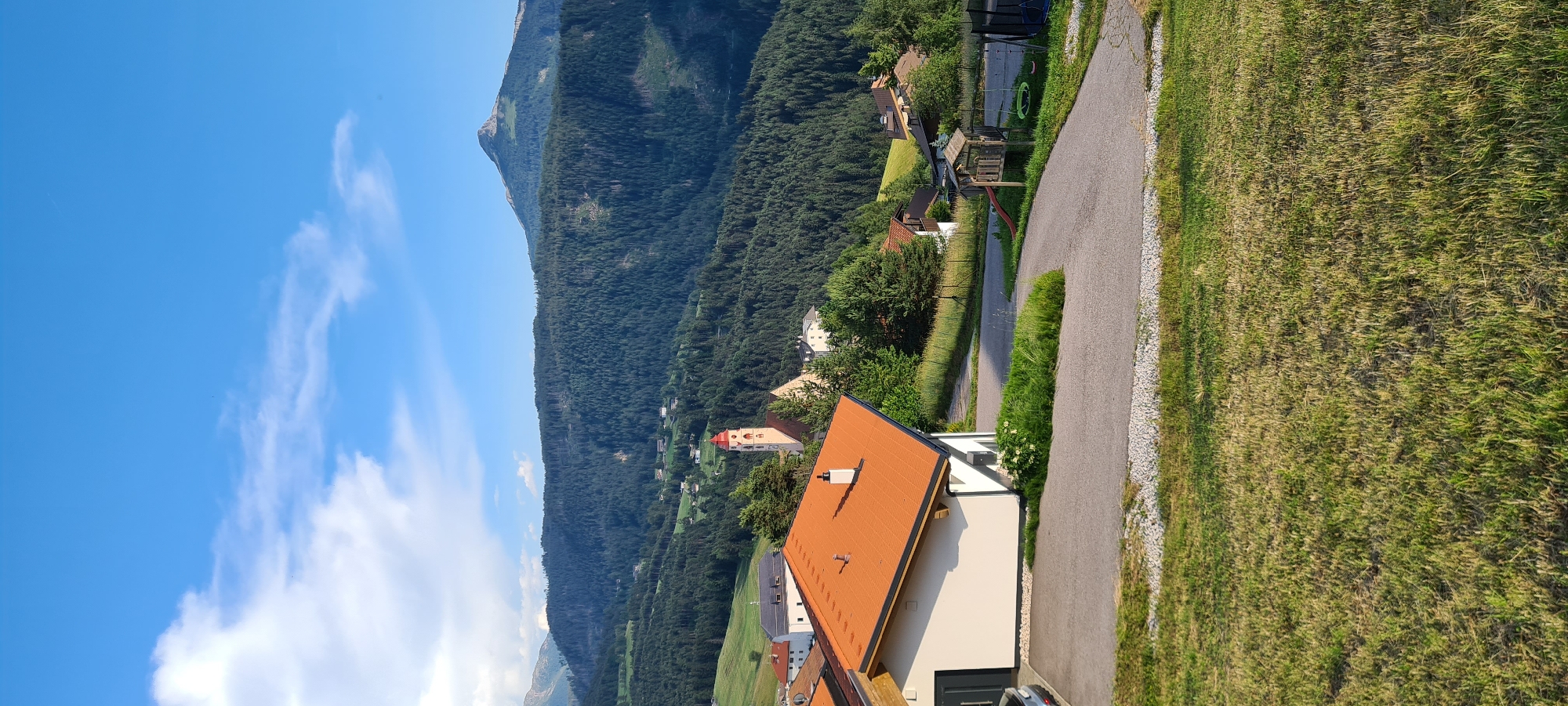 The image captures a scenic view of Deutschnofen - Nova Ponente, Italy, dominated by lush green hillsides and a backdrop of majestic mountains. In the foreground, a modern house with an orange tiled roof is visible. Adjacent to the house is a paved driveway leading to a road that curves through the landscape. A small playground with a swing set and a slide is situated near the road.
Further into the scene, the picturesque village of Deutschnofen unfolds. A prominent church with a red-roofed steeple stands as a central landmark, surrounded by other buildings with traditional architecture. The village is nestled amidst densely forested hills, creating a rich tapestry of green hues that stretch towards the distant, rugged mountain peaks. The sky above is a clear blue, punctuated by a few wispy white clouds, suggesting a pleasant day. The overall impression is one of tranquility and natural beauty, characteristic of the South Tyrolean alpine region.