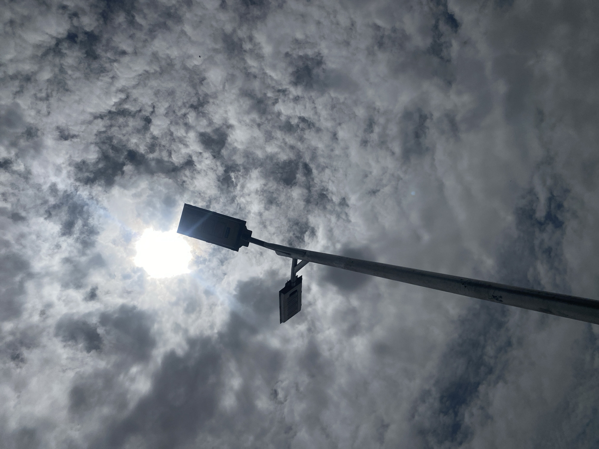 The image shows a low-angle view of a street light against a cloudy sky. The sun is partially obscured by the clouds, creating a bright glare behind the street light. The street light has a pole extending diagonally across the frame from lower right to upper left. There are two light fixtures, one larger and one smaller, attached to the pole. The clouds are dark and textured, filling the majority of the frame. Location is unavailable.