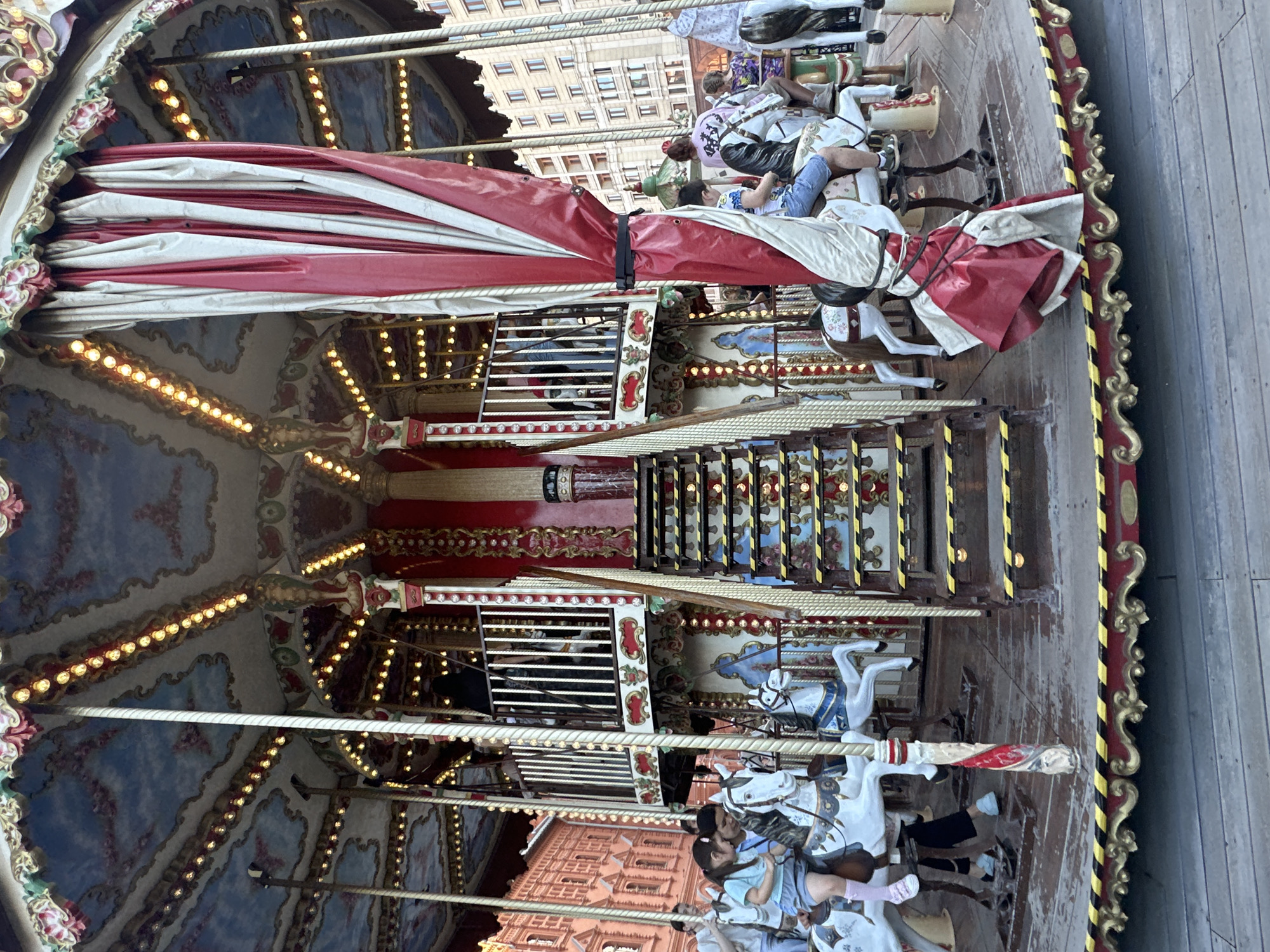 The image depicts a carousel in a city square. The carousel is elaborately decorated with red and white curtains, gold trim, and numerous lights. Several people, including children and adults, are riding on carousel horses. In the background, there are buildings that appear to be of historical significance, with architectural details such as arched windows and decorative facades. The scene suggests a public space within a city, possibly a park or a central square, where families are enjoying a recreational activity. The overall atmosphere is festive and lively. The specific location is Red Square in Moscow, Russia.