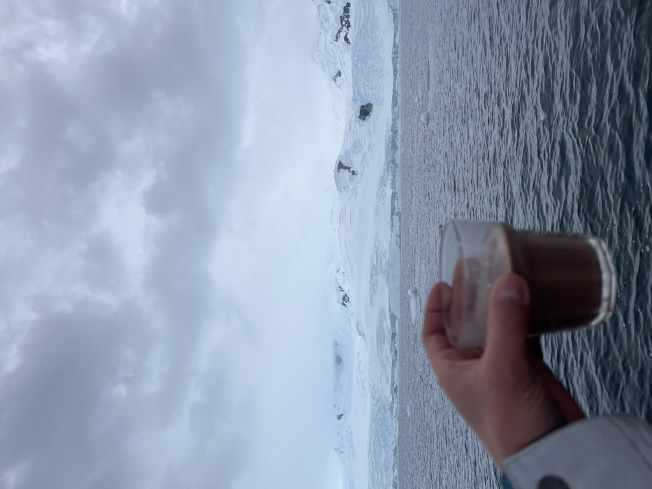 The image presents a serene, cold-weather landscape viewed from a slightly elevated perspective, possibly from a boat or ship. In the bottom right foreground, a person's hand holds a clear glass filled with a dark brown beverage, likely hot chocolate or coffee, about two-thirds full.
Beyond the hand, a vast body of water, appearing as a sea or ocean, occupies the mid-ground on the right, displaying gentle ripples on its surface. Stretching across the mid-ground and background are majestic, snow-covered mountains and glaciers, some revealing dark, craggy rock outcrops. The extensive ice and snow suggest a polar or high-latitude mountainous environment.
The sky, covering the left and upper portions of the image, is heavily overcast with thick, grey clouds, indicating a cloudy or wintery day. The lighting is diffused and soft, creating a muted and tranquil atmosphere. There is no discernible text. The scene evokes feelings of quiet contemplation and the comforting warmth of the drink contrasting with the stark, cold beauty of the natural surroundings.