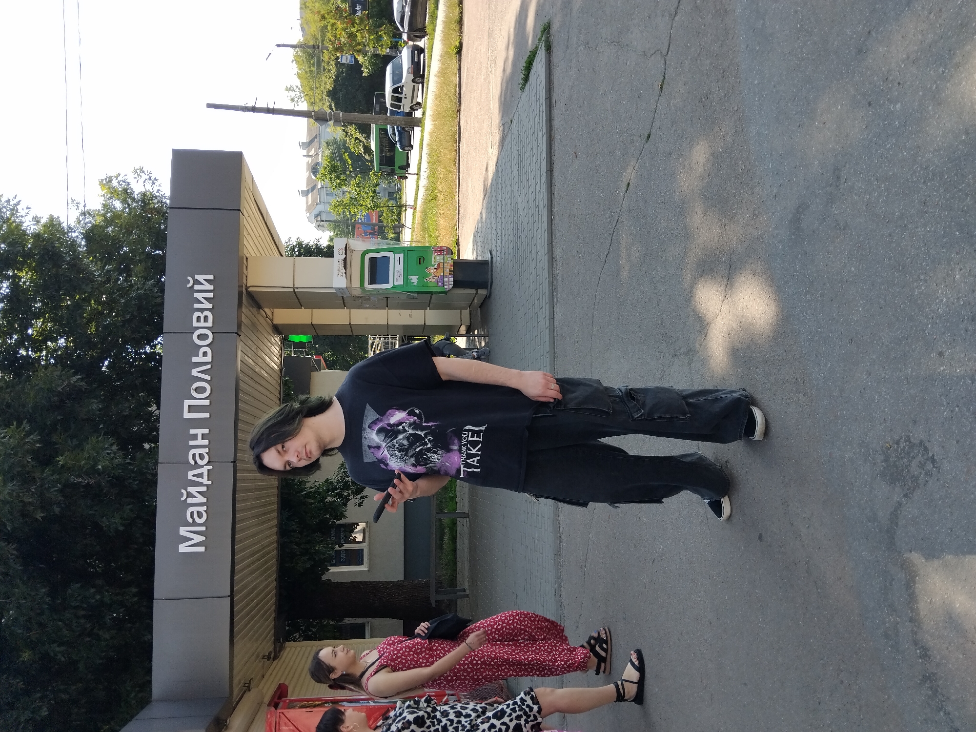 The image depicts a street scene in Zhytomyr, Ukraine, specifically at the Maidan Polyovyi bus stop. The bus stop features a modern grey canopy with white lettering that reads "Майдан Польовий". In the foreground, a young person with long dark hair and green streaks stands holding a smartphone. They are wearing a black t-shirt with a graphic print and dark, baggy cargo pants. To the left, two women are visible. One woman is wearing a black and white leopard print dress and the other is wearing a red jumpsuit with a floral pattern and black sandals. Behind them, a green public transport ticket machine is mounted on a white column. In the background, trees line the street, and a green bus and a white car are visible on the road. The ground is paved with grey interlocking tiles and asphalt. Sunlight casts shadows from the trees onto the ground.