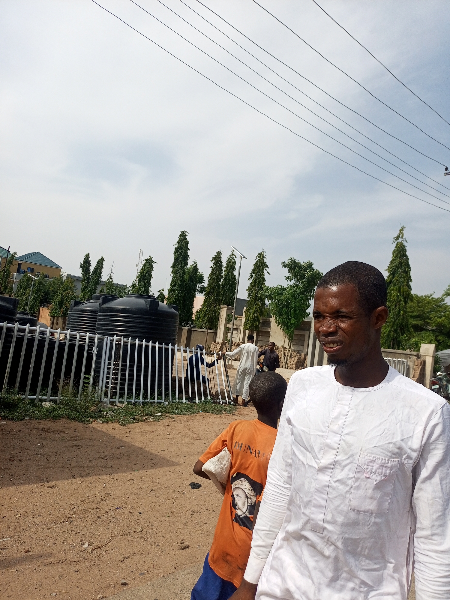This image depicts an outdoor scene with several people, objects, and a clear sky. In the foreground, a man wearing a white traditional garment with subtle patterns and a chest pocket is the primary subject. He has dark skin and short hair, and his facial expression is slightly contorted, possibly due to the bright sunlight. To his left and slightly behind him, another person, a child, is visible wearing an orange t-shirt with a graphic of a historical figure and some text.
The background reveals a row of large black water tanks, presumably for storage. These tanks are behind a white metal fence. Beyond the fence, several tall, slender trees are planted in rows, creating a somewhat formal landscape. In the distance, buildings with light-colored facades and some greenery can be seen. There are also power lines stretching across the upper portion of the image.
Several other individuals are present in the mid-ground, appearing to be engaged in various activities. One person in a white robe is walking, while another in dark clothing is near a motorcycle. The overall setting suggests an urban or semi-urban environment during daytime, with a bright, likely sunny, sky. The presence of water tanks and the outdoor setting could imply a commercial or residential area with a need for water storage. The ground is covered in reddish-brown dirt with scattered debris. The text on the child's shirt is partially visible, reading "DUNAM" above an image.