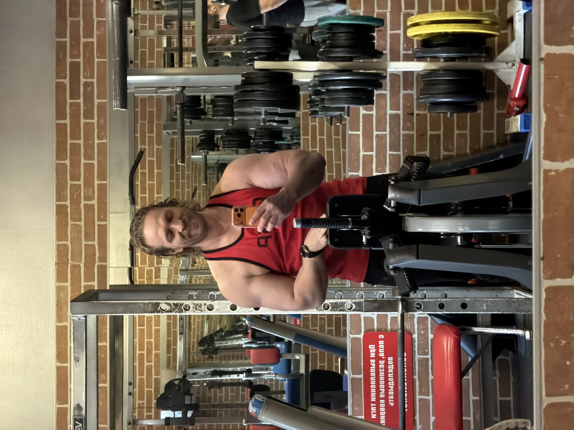A fair-skinned man with long brown hair and a goatee is standing in what appears to be a gym. He is wearing a red tank top with black trim and appears to be taking a selfie with his phone. He is standing near a weight machine. The gym has exposed brick walls and a mirrored wall reflecting weights and gym equipment. The gym appears to be located in Saint Petersburg, Russia, as indicated by a sign in Russian on the weight machine.