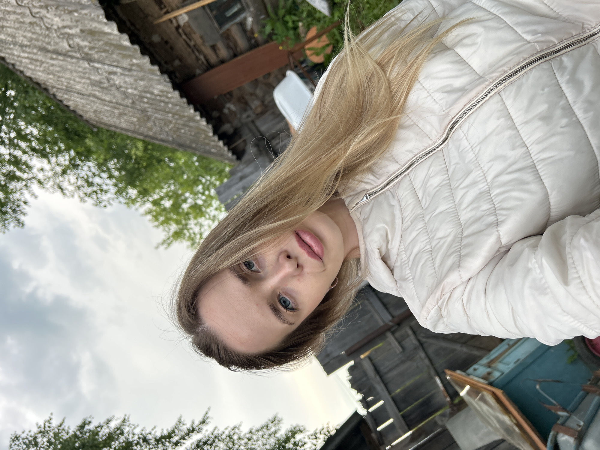 A young woman with blonde hair and blue eyes, wearing a white puffer jacket, takes a selfie outdoors. She is in a rural setting, with a wooden fence and a rustic building in the background. The sky is overcast, and there are trees visible. The location appears to be a backyard or a yard in a village. The image was taken in the city of Азинский, Russia.