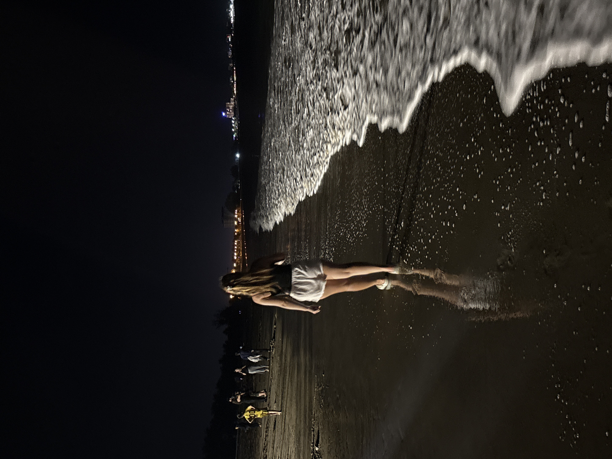 The image captures a night scene on a beach, featuring a woman walking at the water's edge. The woman, viewed from behind, has long blonde hair and is wearing white shorts and a dark top, with her lower legs in the shallow, foamy water. The waves are actively breaking and receding, creating white, frothy patterns on the dark, wet sand.
Further down the beach, several other people are visible, some standing and one appearing to be in a yellow top, possibly kneeling or sitting. The sky is completely dark, confirming the nighttime setting. Along the distant horizon, a prominent line of artificial lights from a city or town illuminates the coastline. The scene depicts a tranquil moment on a beach at night in Dong Hai Ward, Vietnam. There is no visible text in the image.