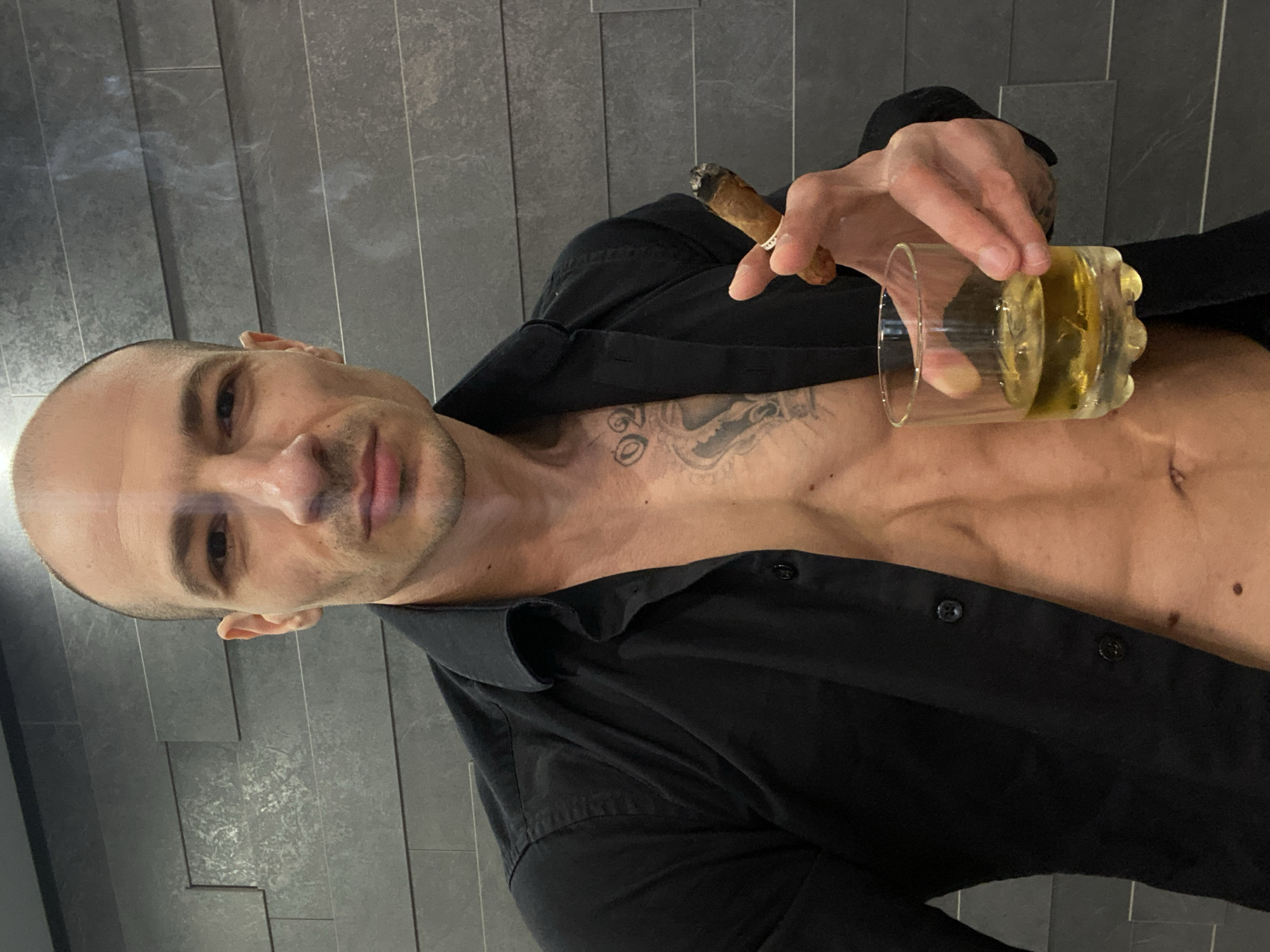 A bald man with light skin stands in front of a gray tiled wall. He holds a lit cigar in one hand and a glass of amber liquid in the other. He wears a black button-up shirt, partially unbuttoned to expose his chest, which features a tattoo. The photo's location and connection to Pleven, Bulgaria, are not discernible from the image.