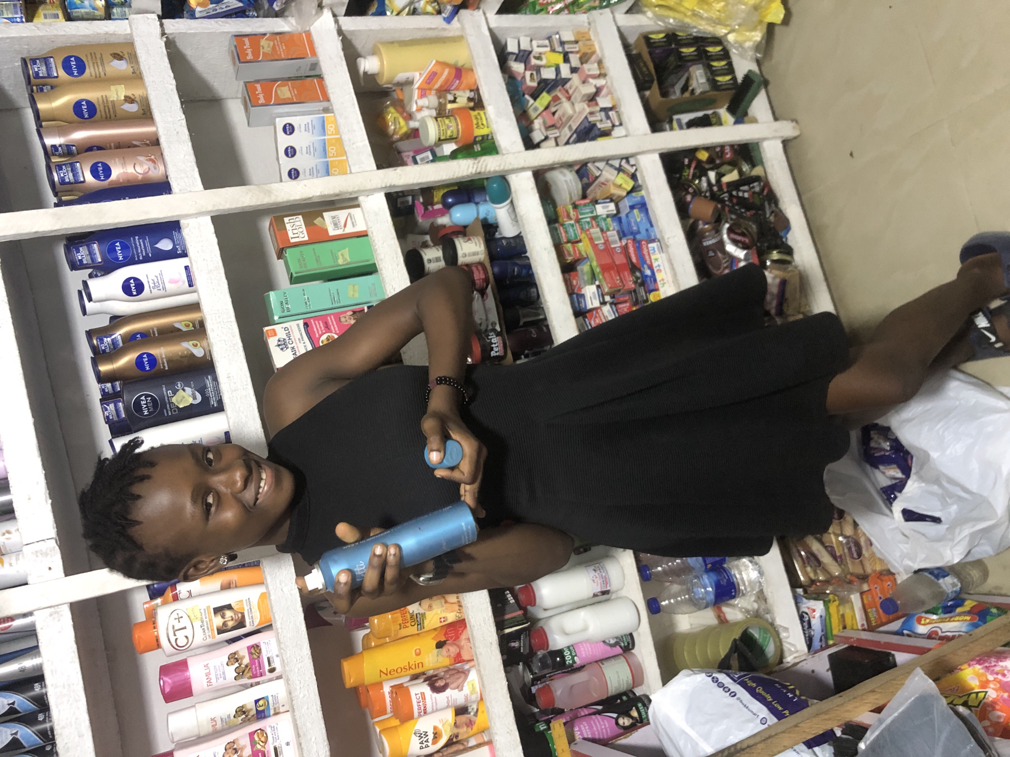 The image shows a young woman standing in a small store that appears to sell toiletries and personal care products. She is wearing a black sleeveless dress and dark blue slide sandals. In her right hand, she holds a blue aerosol can, possibly hairspray or body spray, and in her left hand, she holds a small blue cap, which is likely from the can. Her hair is styled in short, twisted locks. She is smiling and looking directly at the camera.
The store has shelves filled with various products. On the shelves behind her, there are bottles of lotions, soaps, and other beauty products, many of which are Nivea brand. There are also boxes of toothpaste and other small items. The shelves are made of white wood. The floor appears to be tiled in a light color. The overall impression is that of a local convenience store or pharmacy. This scene could be in Ado, Nigeria, as the prompt suggests.