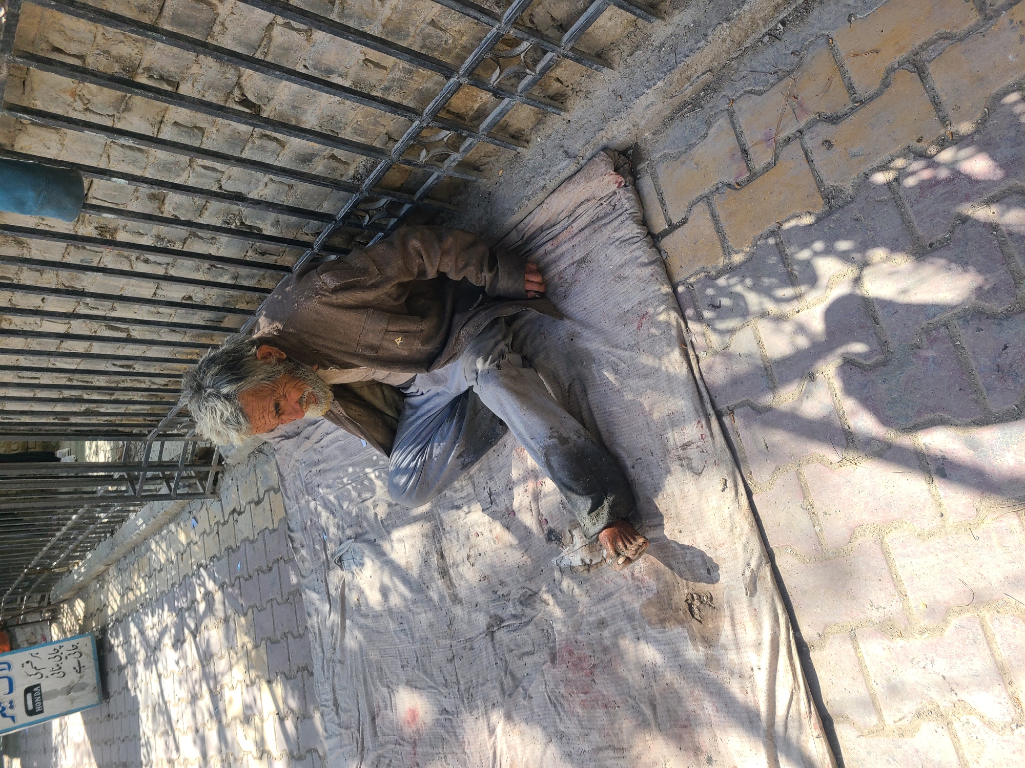 An elderly man with grey hair lies on a white cloth on a brick-paved surface next to a metal grate in Quetta City Tehsil, Pakistan. He is wearing a brown jacket and light-colored pants, and his bare foot is visible. Shadows from a nearby object fall across the brick pavement. A blue sign with text is visible in the background.