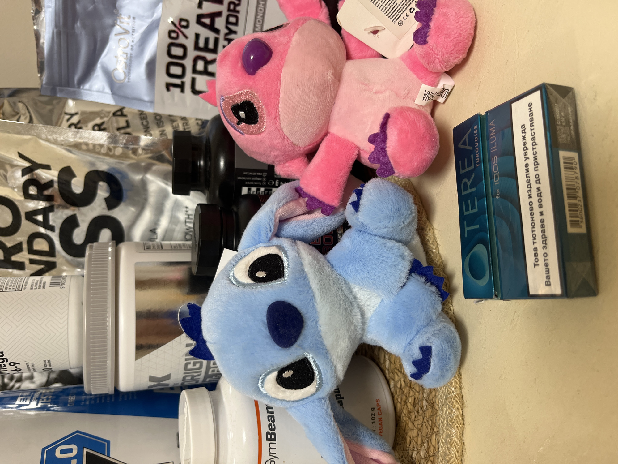 The image shows a collection of items arranged on a surface. A blue and a pink plush toy are positioned alongside supplement bottles and pouches, including a "GymBeam" bottle and a "100% Creatine Hydrate" pouch. There are also packs of "TEREA TURQUOISE for IQOS ILUMA" smoking products. The text on the packs is in Bulgarian, stating "Това тютюнево изделие уврежда Вашето здраве и води до пристрастяване" ("This tobacco product harms your health and leads to addiction"). Given the language, this image is likely from Bulgaria, though the city of Nova Zagora cannot be confirmed from visual data.