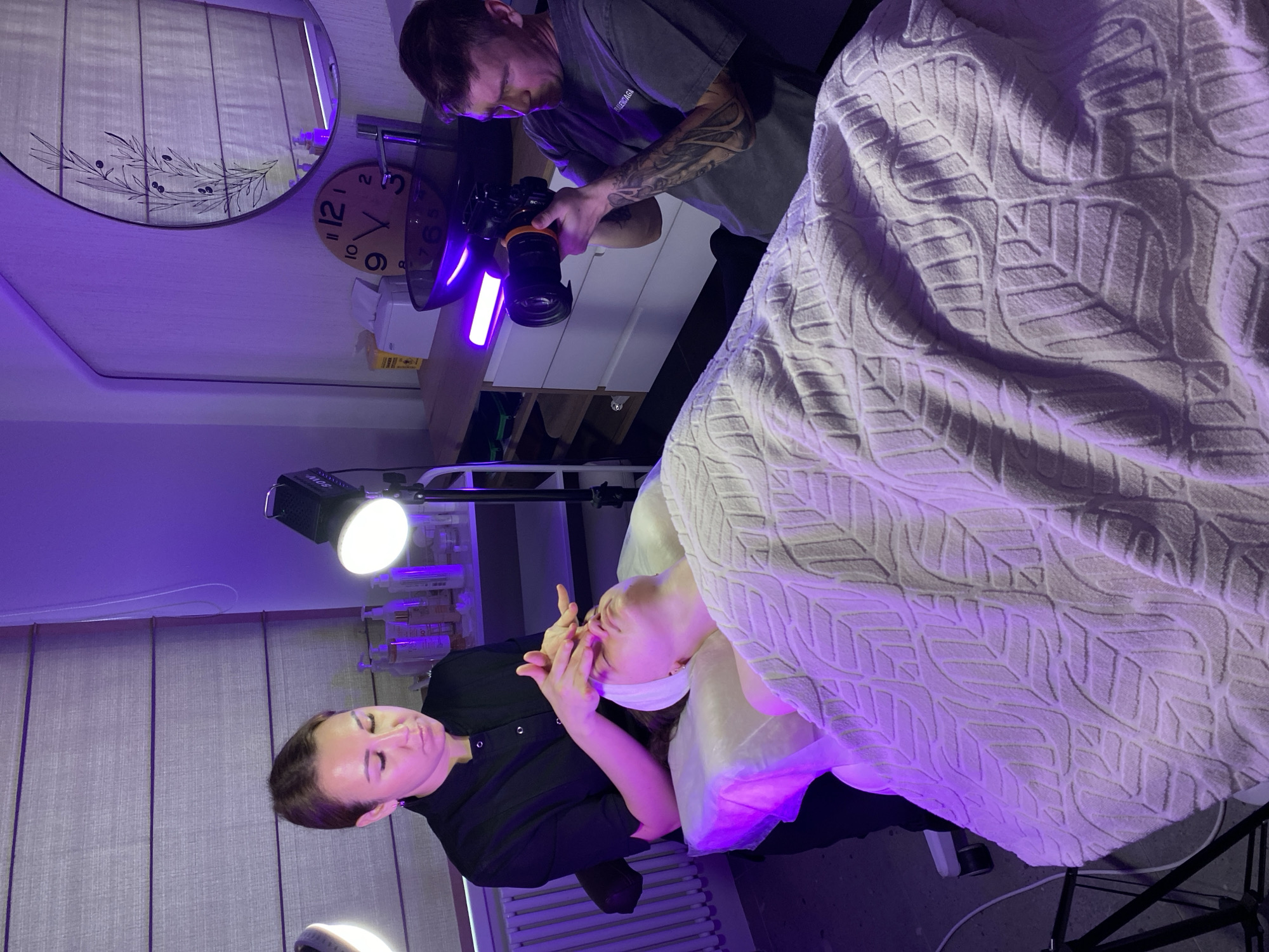 In a room with purple lighting, a woman in a black uniform is performing a facial treatment on a client lying on a treatment bed covered with a patterned white blanket. Another man stands nearby, holding a camera and taking photos. Background details include a clock on the wall, cosmetics on shelves, and a window with blinds. The specific location in Izhevsk, Russia, is not discernible from the image.