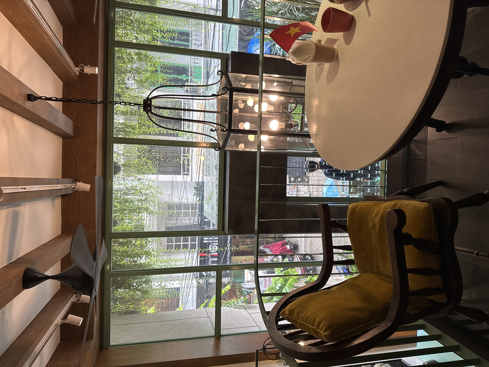 A sun-drenched interior view shows a room with an aged, antique rocking chair, a small round table with an iced beverage and a miniature Vietnamese flag placed on top of it, a black metal framed decorative lantern, and a ceiling fan. The setting appears to be a cafe or restaurant. A large multi-paned window offers a view of a street scene, hinting at an urban environment. Address is written on one of the shops, with street numbers 92/17. The city Nha Trang Ward in Vietnam. The chair is made of dark wood, with golden yellow fabric on the seat and back cushions. The table is round and white with a black metal frame. The lantern is ornate and hangs from the ceiling. The ceiling fan is black and is mounted on the ceiling. The window is large and offers a view of the street outside, which includes pedestrians, shops, and buildings. The street is lined with trees.