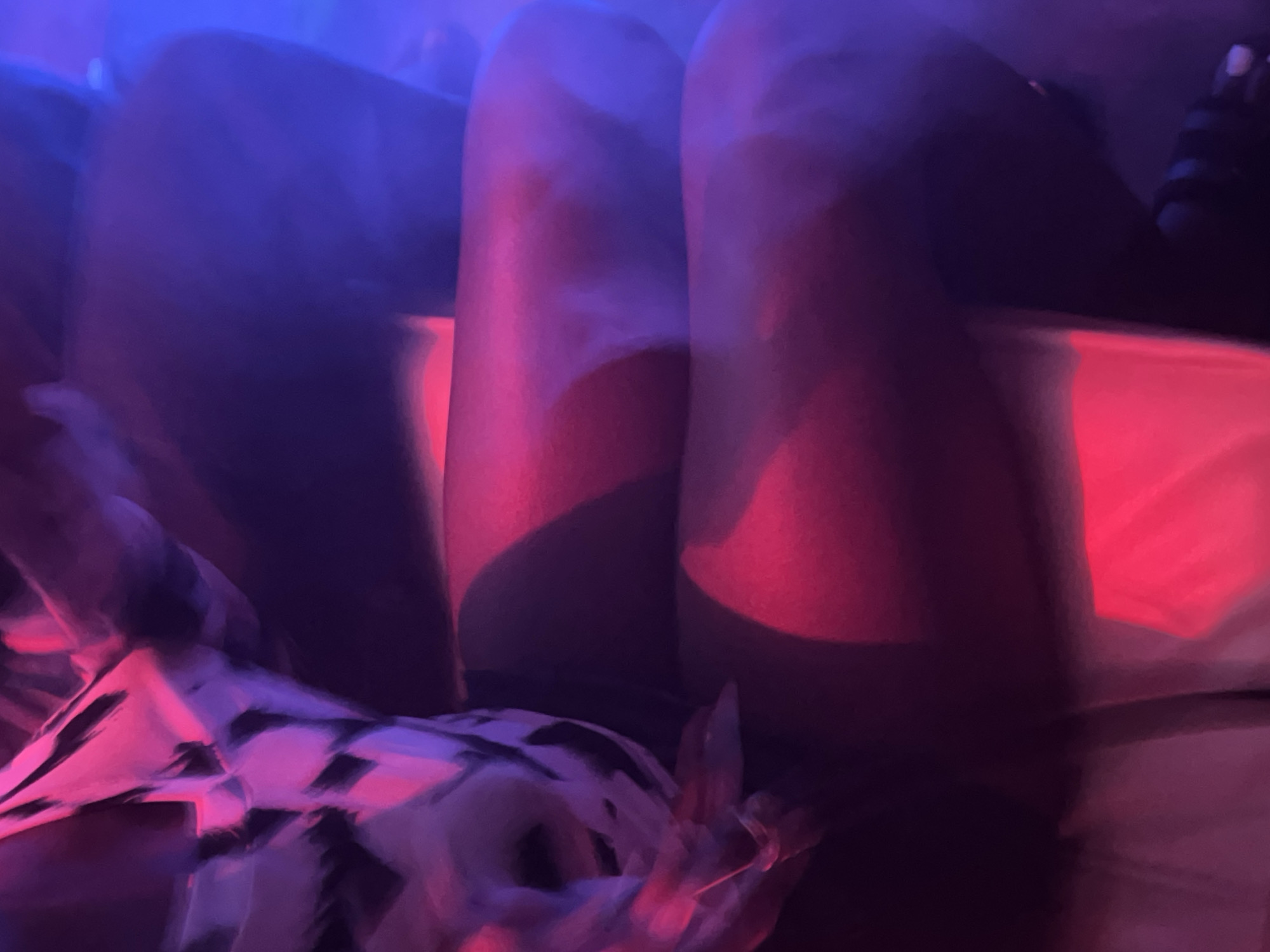 The image shows a close-up view of a person's legs, illuminated by red and blue lighting. The person is wearing a patterned top and dark bottoms. The setting appears to be indoors, with soft shadows and blurred edges suggesting a low-light environment, possibly a club or a private gathering. There is no indication of the location being in Gura Top, Nigeria.
