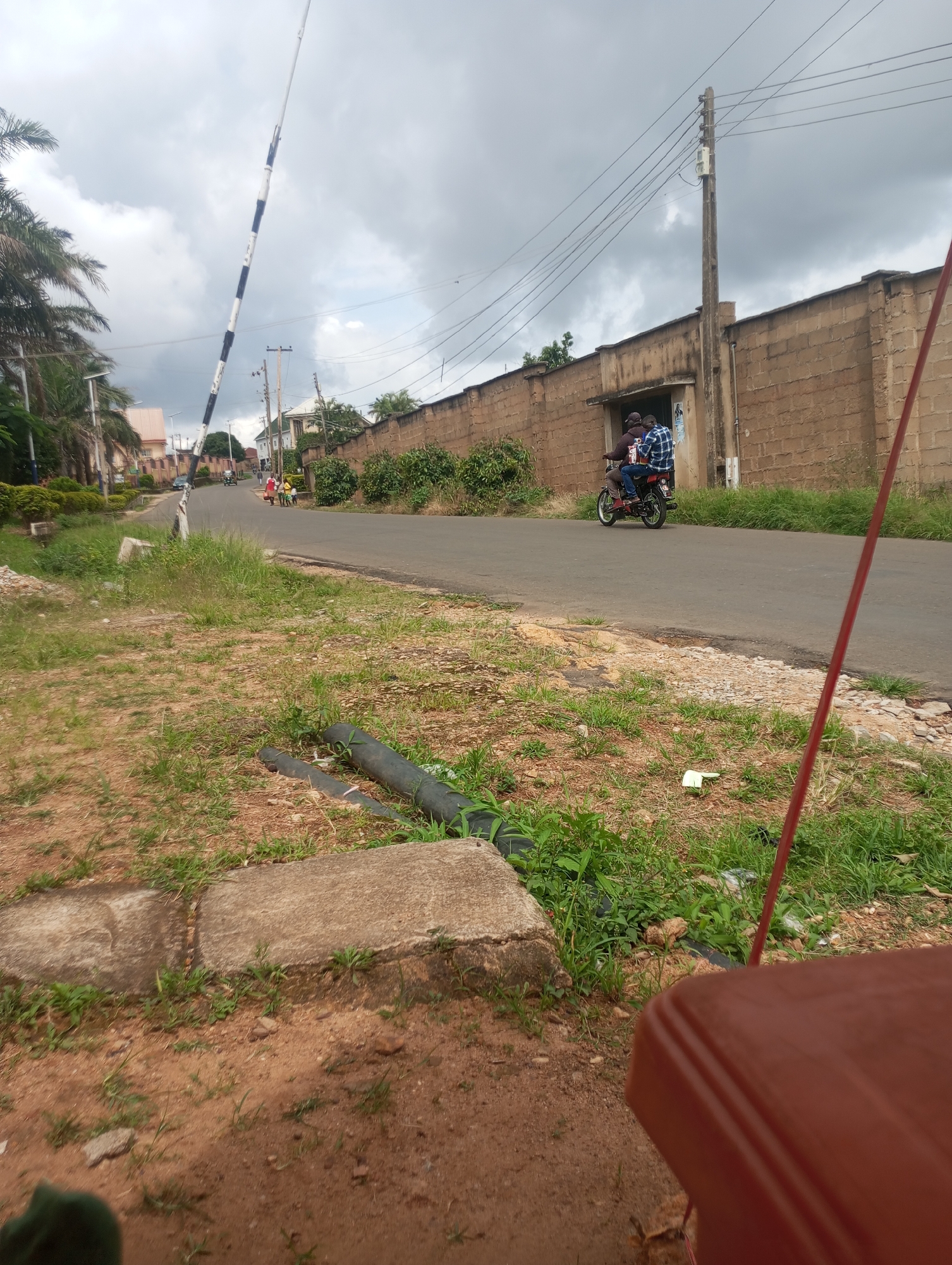 In the city of Jos, Nigeria, a cloudy sky looms over a street scene. A tall, black and white striped barrier, possibly a railway crossing gate, stands at an angle on the left side of the image. A paved road stretches into the distance, lined with buildings and lush green vegetation.
On the right side, a brick wall defines the edge of a property, with a tall utility pole and overhead wires running parallel to it. A motorcycle carrying two individuals, one in a plaid shirt, travels down the road. Further down the road, a few people are visible walking.
The foreground of the image is dominated by dry, uneven terrain with patches of grass and some concrete structures. A dark, flexible pipe lies on the ground, and a red pole is visible extending upwards from the bottom right corner of the frame. The overall impression is of a somewhat unkempt, semi-urban environment under an overcast sky.