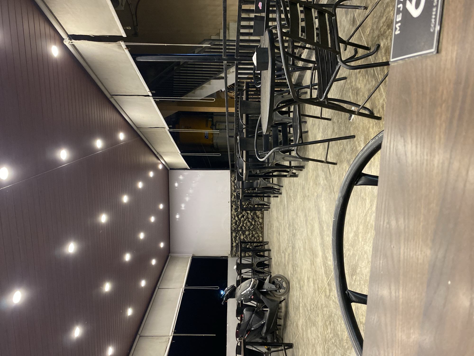 The image depicts an outdoor seating area, likely a café or restaurant, in Keudeublang, Indonesia. A row of black metal chairs and tables are arranged on a concrete floor, with some chairs stacked. A black motorcycle is parked in the seating area. Above, a brown ceiling with recessed lighting extends over the space. A sign with the word "MEJA" is visible on one of the tables.