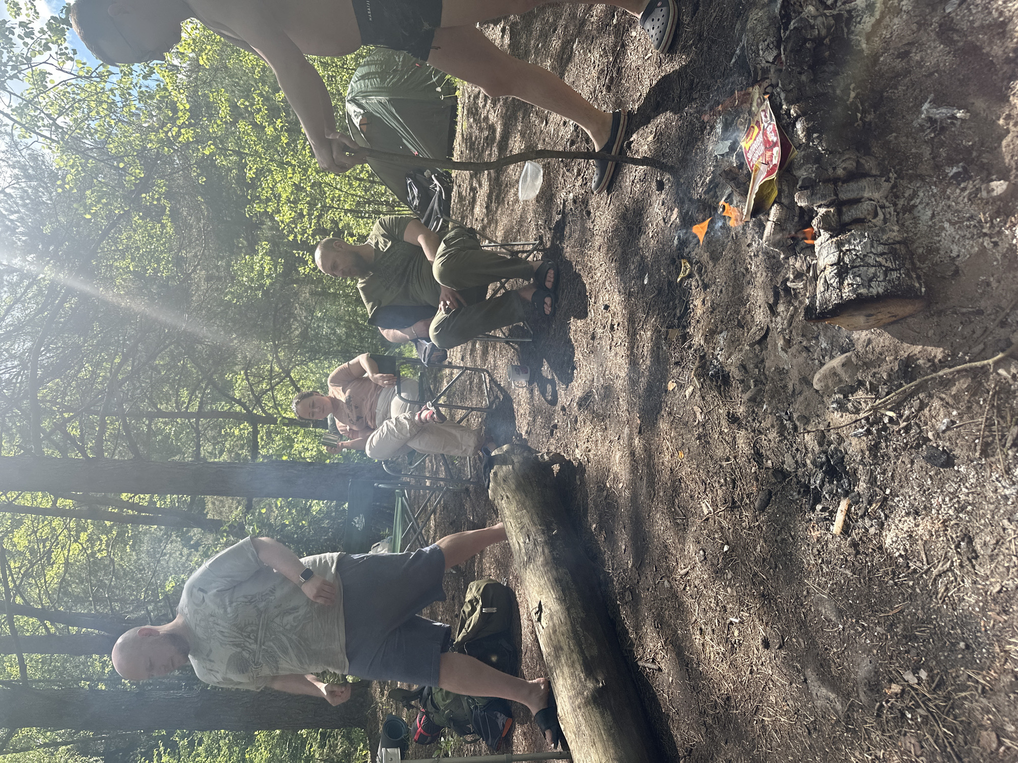 The image shows a campsite in a wooded area, likely in Юзуфоўскі сельскі Савет, Belarus. Four people are visible, sitting in camping chairs around a small campfire. One person stands close to the fire holding a stick. A tent is set up nearby. The ground is covered with dry leaves and dirt. A fallen log lies close to the campfire. There are backpacks, other camping equipment, and trash visible around the campsite.