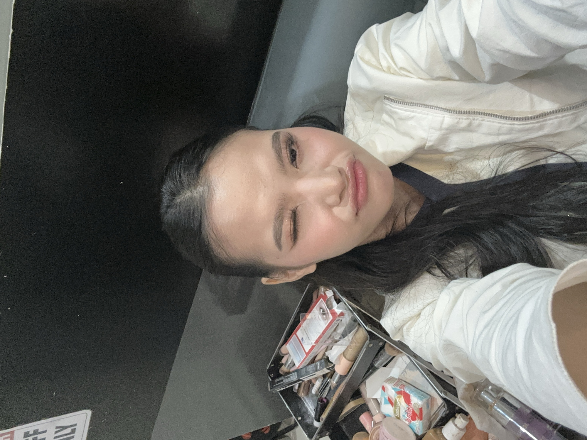A young Asian woman is captured in a close-up, selfie-style shot, appearing to be in a backstage or dressing room environment, possibly in Davao City, Philippines. She is looking directly at the camera with her left eye closed in a wink, and her lips are pursed. She has on makeup, including eyeshadow and mascara, and her hair is dark and pulled back. She is wearing a light-colored jacket over a dark garment. To her left, a large, open makeup case is visible, filled with various cosmetics, brushes, and applicators. The background is dark and appears to be a wall or partition. A sign with partial text, possibly "FF ILY," is visible in the upper left corner. The lighting suggests an indoor setting.