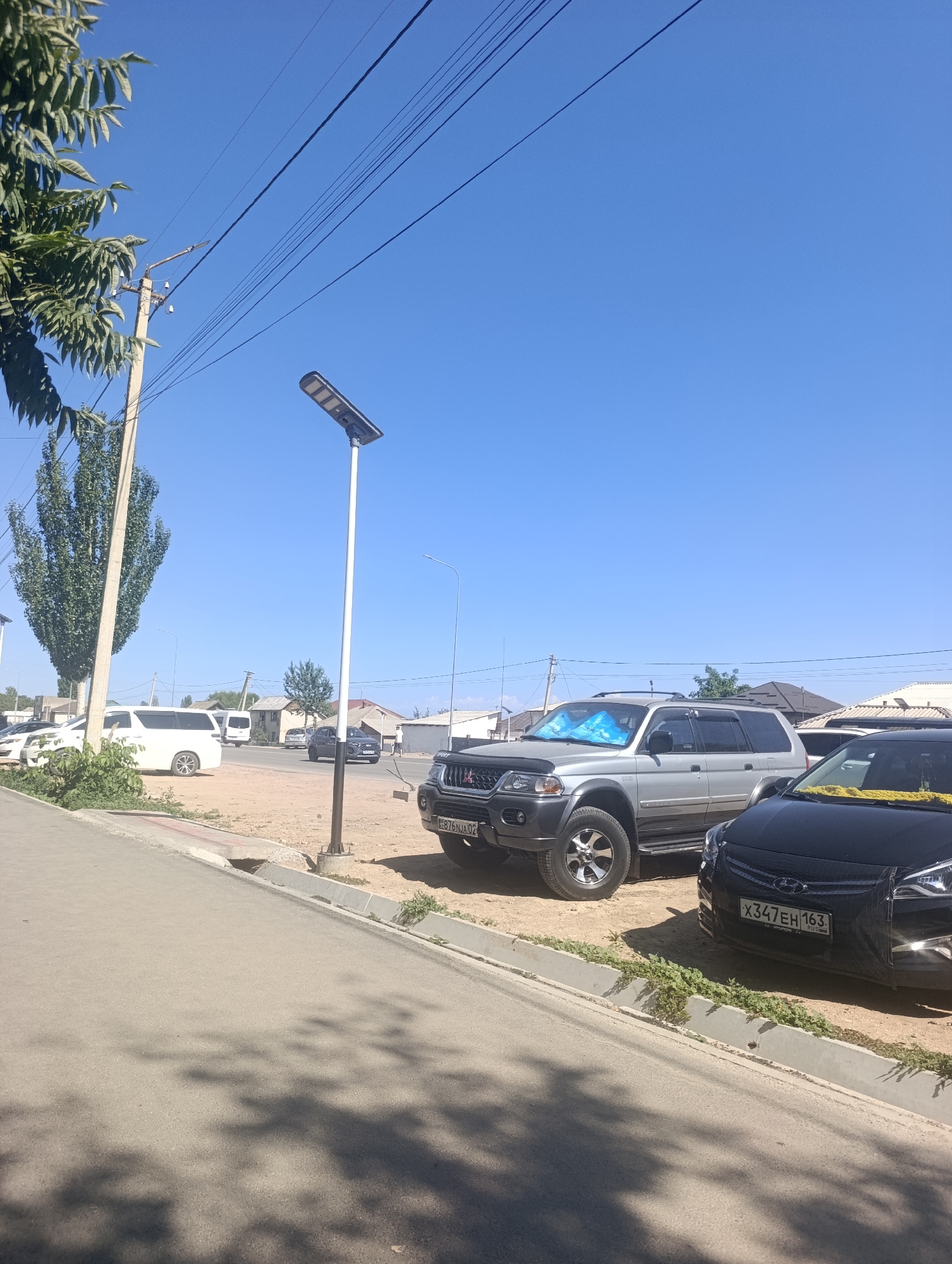 The image shows a street scene in Bosteri, Kyrgyzstan, under a clear blue sky. On the left, a paved road runs parallel to a raised sidewalk with a concrete curb. Beyond the sidewalk, there is a patch of dry, sandy ground with sparse vegetation and scattered debris. A tall, slender tree with dense green foliage stands prominently on the left side of the frame, adjacent to a utility pole and overhead power lines.
Several vehicles are parked or driving on the road. A white van is parked on the left side of the road, and further down, a dark-colored SUV is visible. A silver Mitsubishi Pajero SUV is parked in the center of the frame, with a blue sunshade visible on its windshield. To the right of the silver SUV is a black Hyundai sedan. A license plate on the black Hyundai reads "X347EH 163". A man is standing near the sidewalk on the right side of the road. In the background, residential buildings with pitched roofs are visible, along with more trees and utility poles. The overall impression is of a sunny day in a semi-urban or rural area.