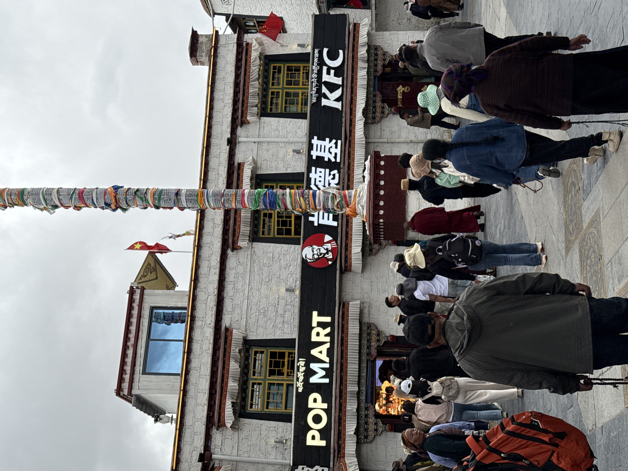 The image shows a building in the Chengguan District, Lhasa, Tibet, China, with a KFC restaurant identified by a black sign displaying "KFC" and Chinese characters. Below it, another sign reads "POP MART" in large white letters. Prayer flags are strung across the building's facade. A crowd of people is present in front of the building, some of whom are prostrating themselves on the ground, possibly in prayer. The building has a traditional Tibetan architectural style. The sky is overcast.