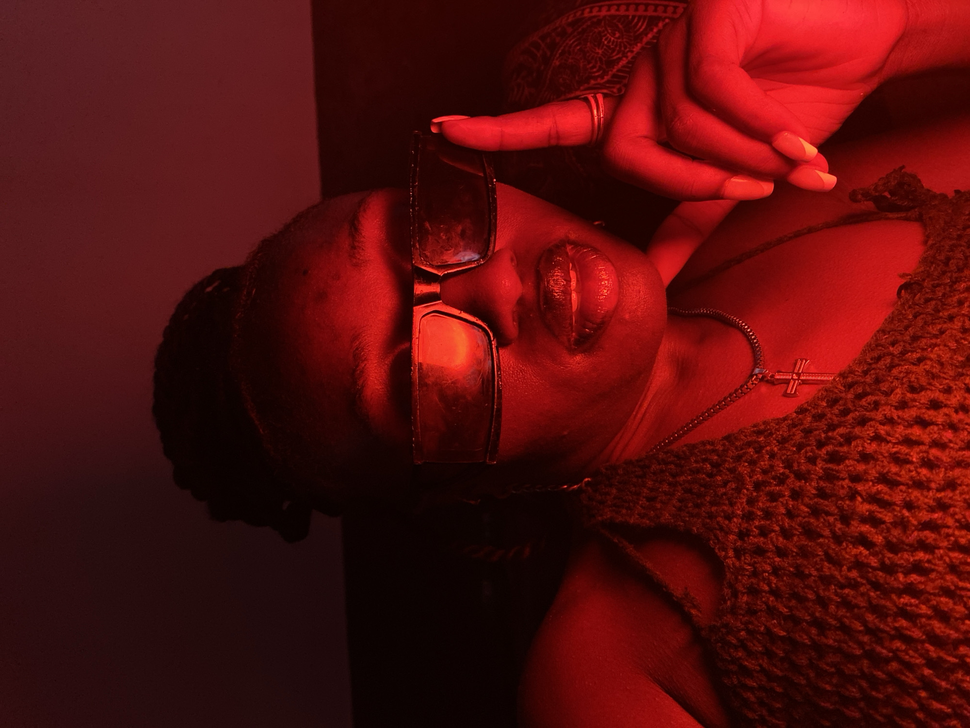 This is a close-up, vertically oriented, low-angle shot of a person, illuminated by red light. The subject is lying down, with their head tilted back and their eyes closed. They are wearing large, dark-rimmed glasses that are being held up by their right hand. The hand has manicured nails painted with a light color, and the subject is wearing rings on two fingers.
The subject has full lips that appear to be glossy or moisturized. Around their neck, they are wearing a necklace with a small cross pendant. They are also wearing a dark, crochet-like garment that covers their chest and shoulder. The background is dark, with the red light casting a strong hue over the entire scene. The lighting creates a moody and intimate atmosphere. The location context of Bar Jirgi Summa, Nigeria, is not visually evident in the image itself. There is no visible text in the image. The overall impression is one of sensuality and repose.