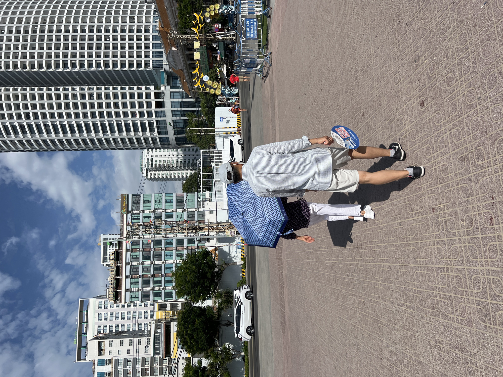 In Nha Trang, Vietnam, two individuals walk across a red brick paved plaza in front of several modern buildings. The person in the foreground, facing away from the camera, wears a striped button-down shirt and light-colored shorts, and carries a circular fan in their right hand. To their left, a second person, also facing away, holds a large blue and white patterned umbrella, obscuring most of their upper body; they are wearing white pants and a dark patterned shirt. The background features a tall, cylindrical white building with numerous balconies, a collection of mid-rise buildings with multiple windows, and a street with a white SUV and other vehicles. The sky is blue with scattered clouds. The scene appears to be a public area on a bright, sunny day, possibly near a waterfront or tourist district given the architecture and open space.