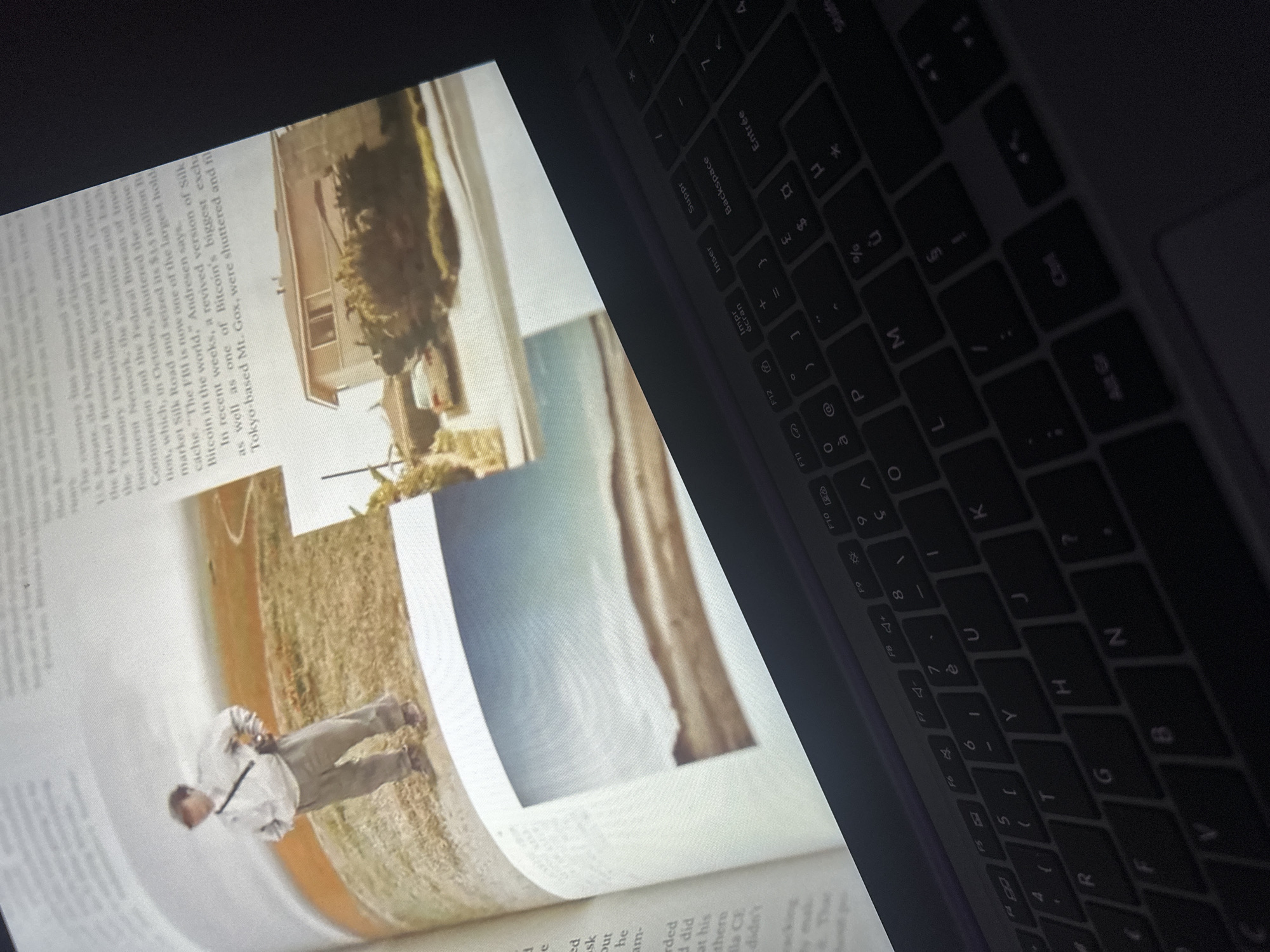The image depicts a close-up view of an open book lying on a dark laptop keyboard. The book features several photographs and text. One photograph shows a person standing in an open, grassy field. Another photograph shows a residential house with a car parked in the driveway. The text in the book appears to be about a financial or technological topic, mentioning "Bitcoin" and "Silk Road". The laptop keyboard is partially visible, with keys like "Entrée", "Backspace", "Insérer", "Suppr", "Alt Gr", and "Ctrl" discernible. The lighting suggests the scene is indoors, possibly at night, with the light from the laptop screen illuminating the book. There is no visual information in the image that indicates the location is in the city of Anduze, France.